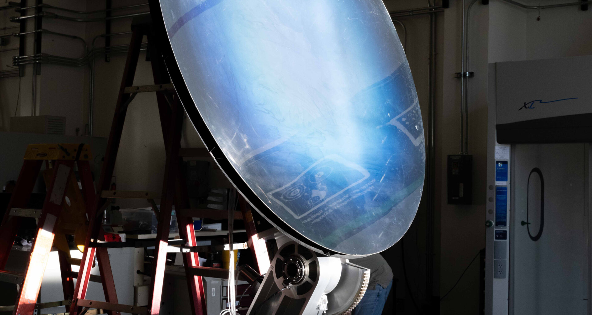 Light shines onto a mirror-like solar concentrator resting on a workbench. The concentrator is tilted upward around 45 degrees to catch the light. Its surface is black at the edges, and as you move inward, it goes from a deep blue to white.