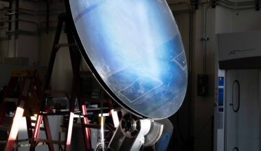 Light shines onto a mirror-like solar concentrator resting on a workbench. The concentrator is tilted upward around 45 degrees to catch the light. Its surface is black at the edges, and as you move inward, it goes from a deep blue to white.