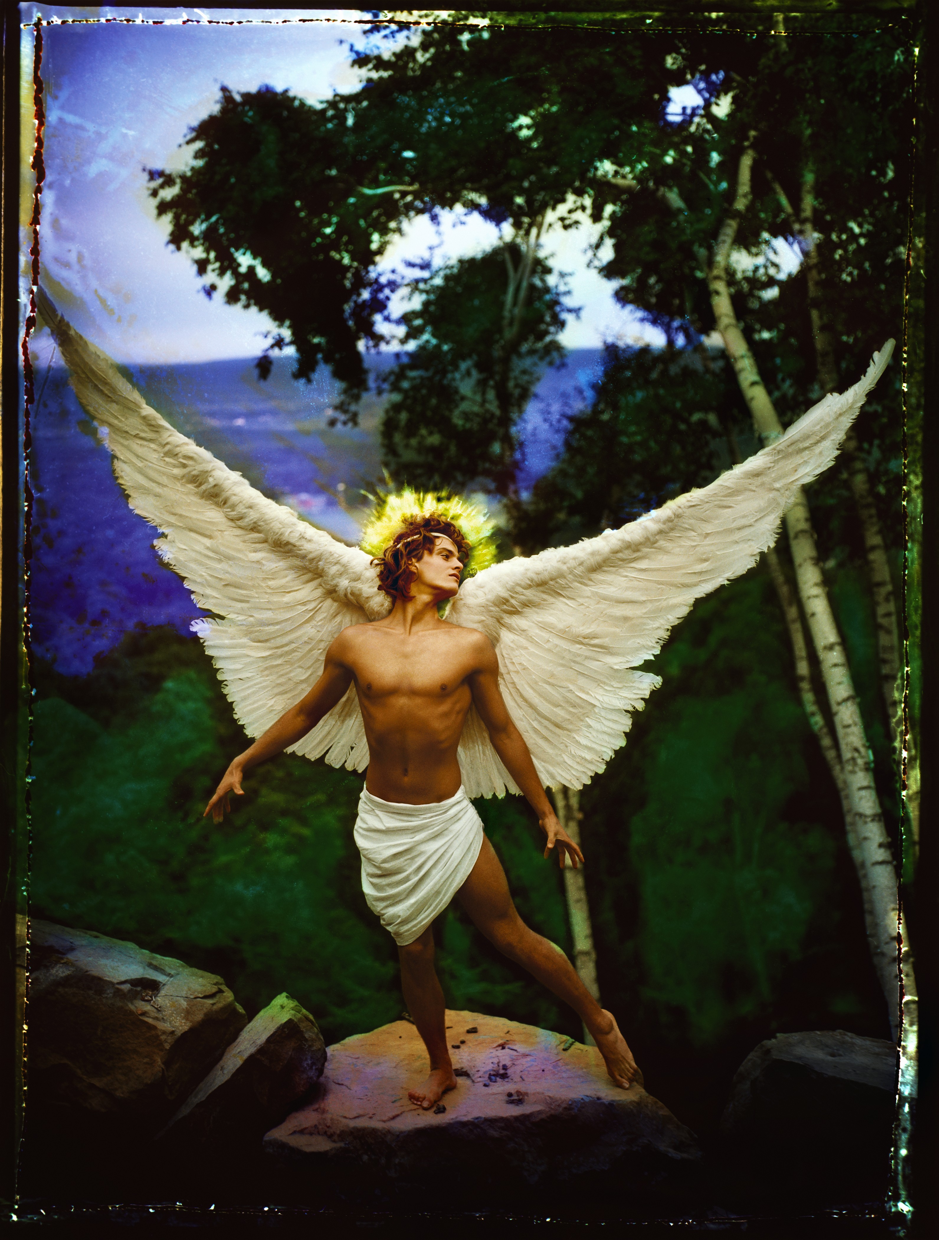 A person with large, white feathered wings and a glowing golden halo stands atop a rock in a lush, green forest setting.