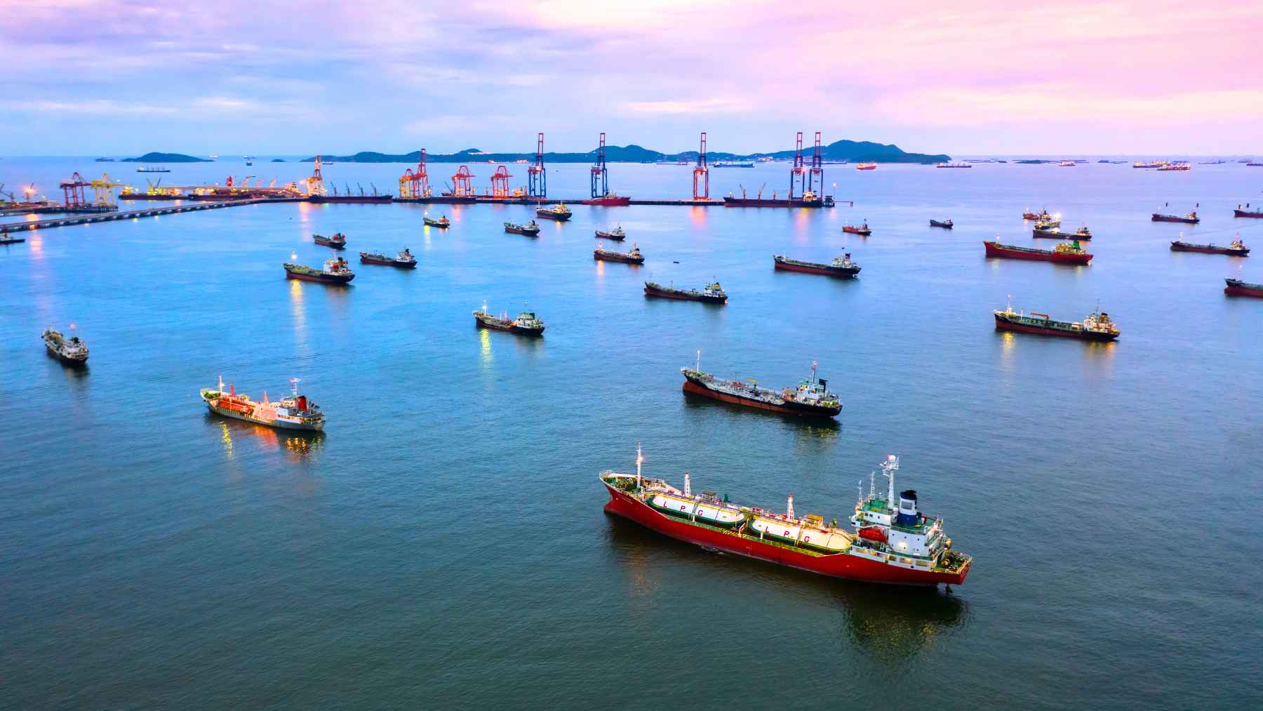 Cargo ships sit offshore near a busy port at dusk, a visual of global shipping and the climate impact of cleaner marine fuels.
