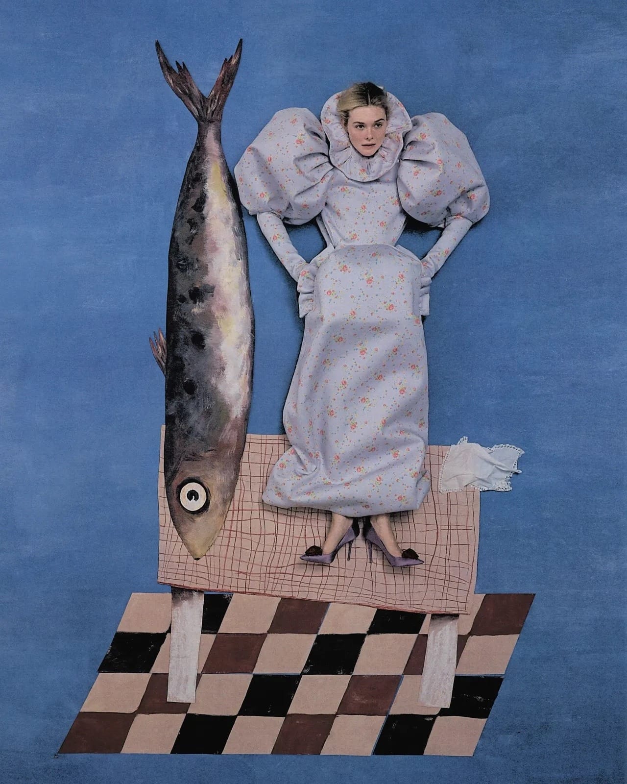 an editorial flat-lay portrait by Szilveszter Makó of Elle Fanning posed next to a giant sardine. She and the fish stand atop a table that sits on a checkered floor.