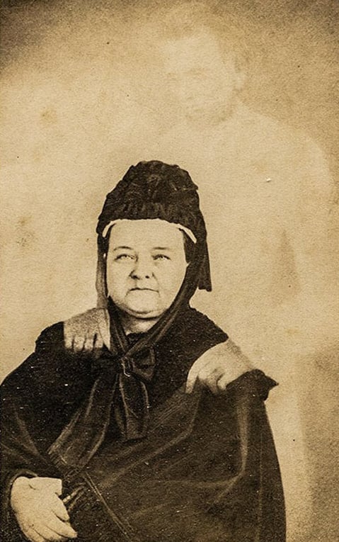 An old sepia-toned photograph of a seated woman in dark clothing and a bonnet, with a faint, ghostly figure of a man standing behind her, his hands resting on her shoulders.
