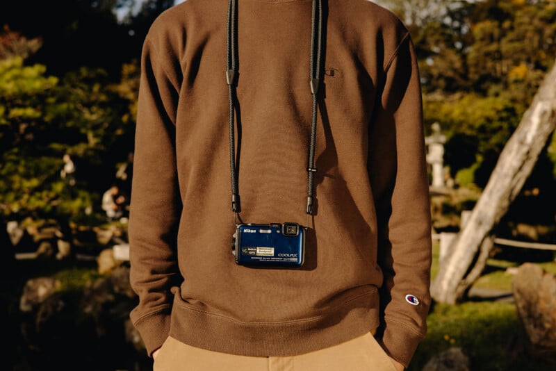 A person wearing a brown sweatshirt and tan pants stands outdoors, with a blue Nikon Coolpix camera hanging from their neck on a black strap. The background is blurred greenery and sunlight.