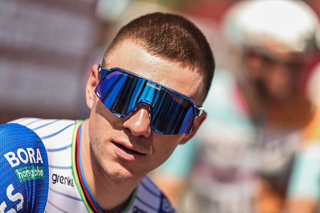 A close-up shot of Remco Evenepoel wearing large sunglasses and wearing the Red Bull-Bora-Hansgrohe kit with rainbow trim on the collar