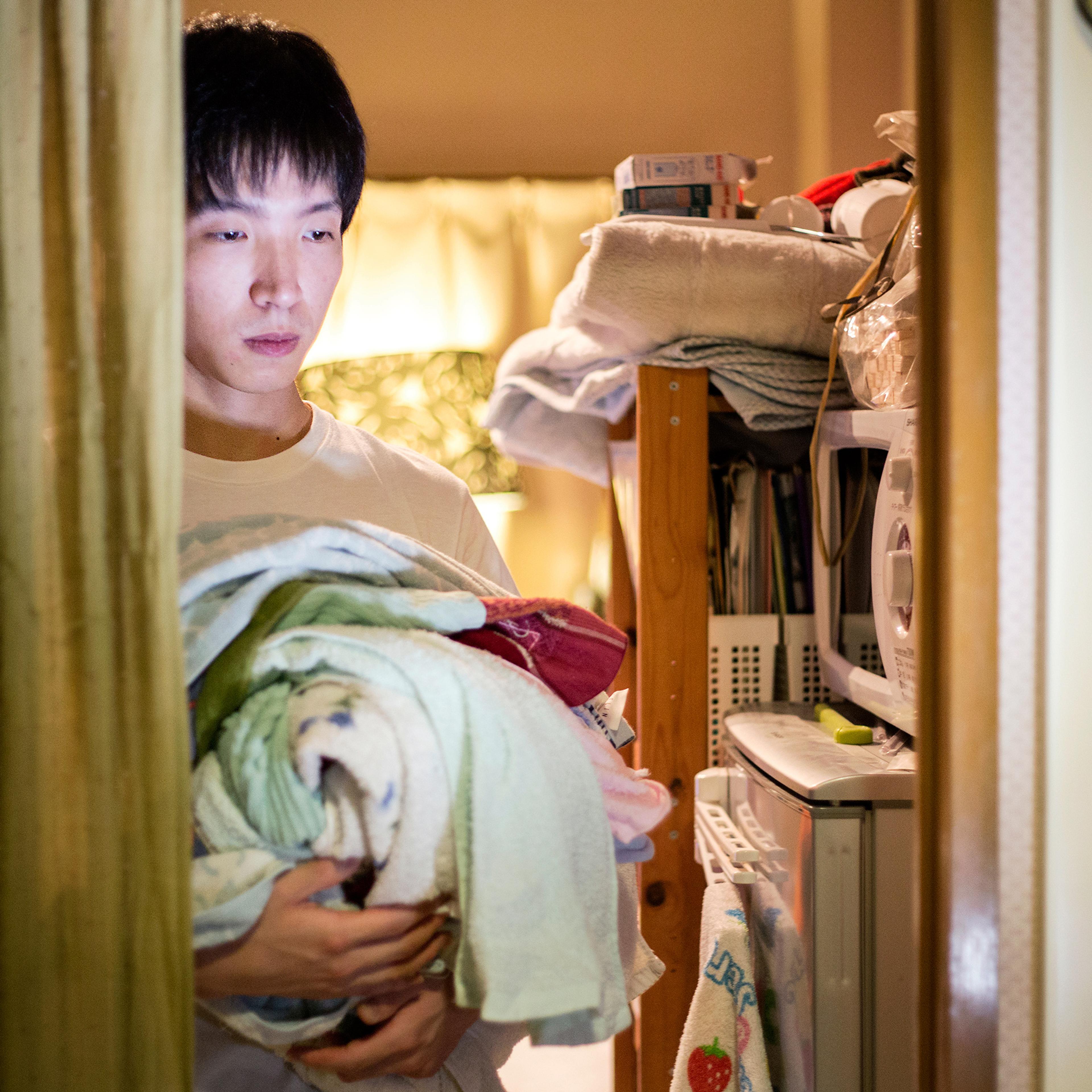 Photo of a man holding laundry in a dimly lit room with shelves, a microwave and a curtain on the left.