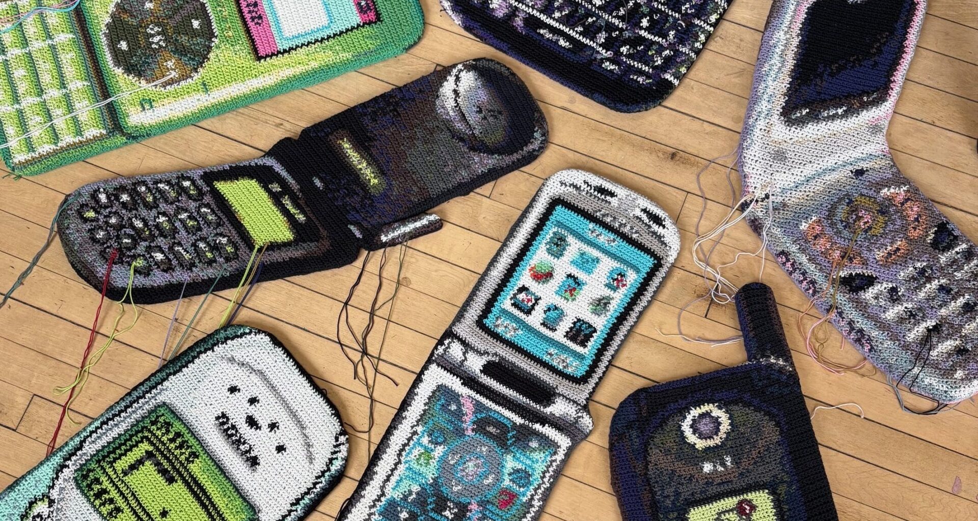 Fiber art pieces by Nicole Nikolich of dated flip phones, laid out on a hardwood floor