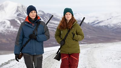 First look from Vigil series three. DCI Amy Silva (Suranne Jones) and DI Kirsten Longacre (Rose Leslie) walk across a snowy landscape. They wear warm winter clothing and carry rifles over their shoulders. 