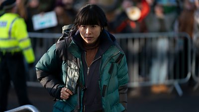 First look from Vigil series three. A woman wearing a heavy winter coat carries a backpack. A police officer in the background watches crowds behind a barricade. 