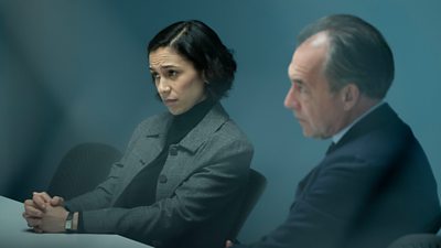 First look from Vigil series three. A woman and a man in formal clothing sit at a table in an interrogation room. 
