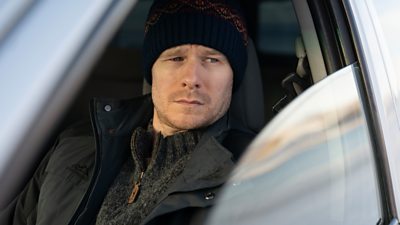 First look from Vigil series three. Close up of a man sat in a car, watching something through the open window. 