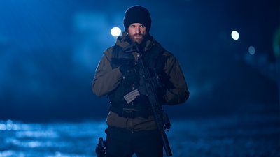 First look from Vigil series three. A man stands outdoors at night carrying a large rifle against his chest. Dressed in warm clothing he watches something offscreen. 