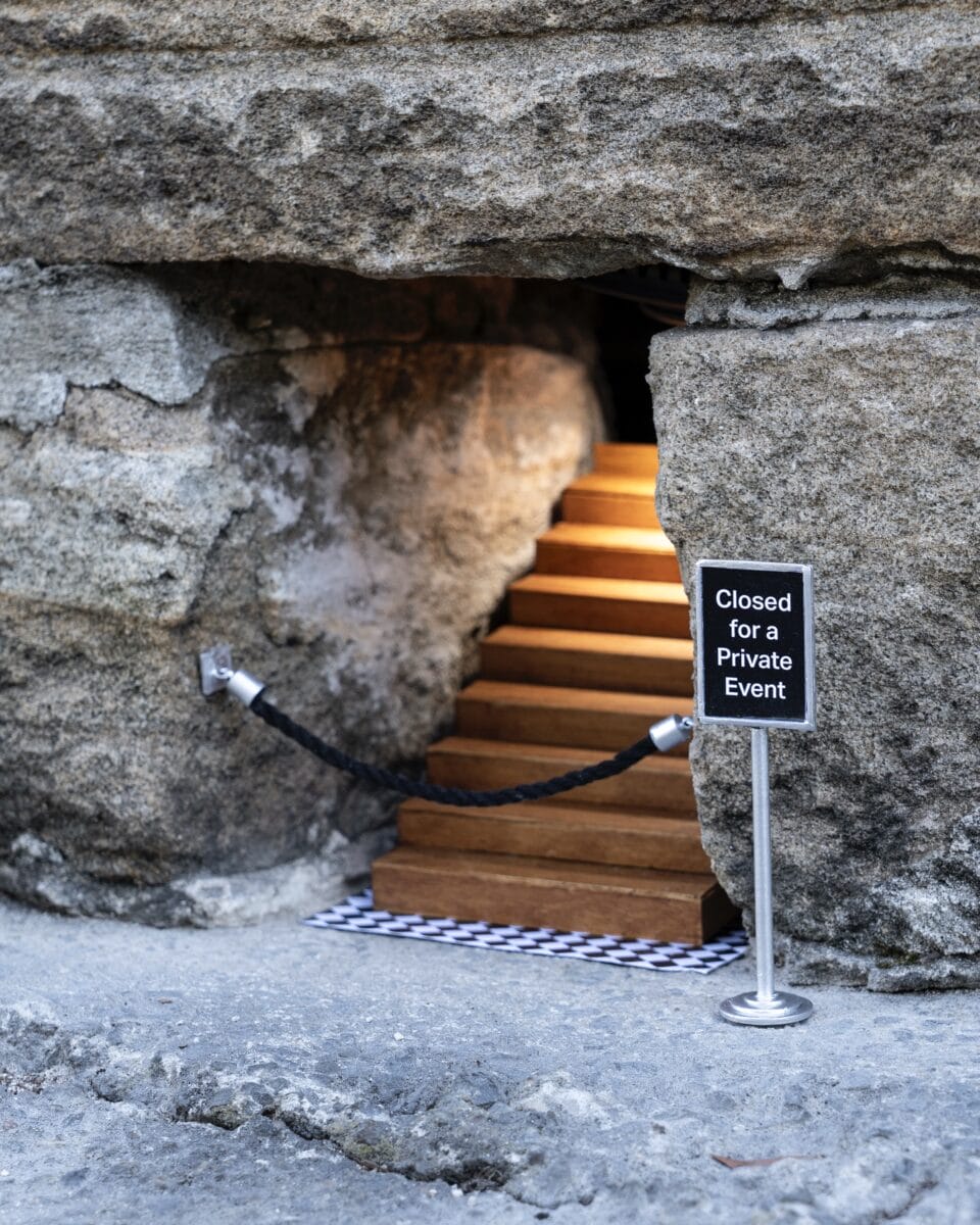 A miniature installation by Michael Pederson of a gap at the base of a stone wall with a tiny stairwell leading up into it and a stanchion with a sign reading