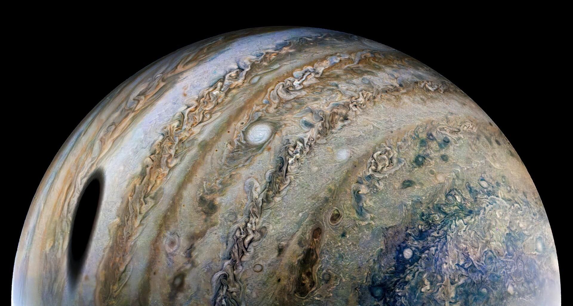 View of one-half of Jupiter. Swirling rings of gold, brown, green, and blue dust and gas clouds wrap vertically around the sphere of the planet. A large dark spot is seen toward the bottom left and smaller spots of white dot the surface, breaking up the rings of swirling clouds and dust.