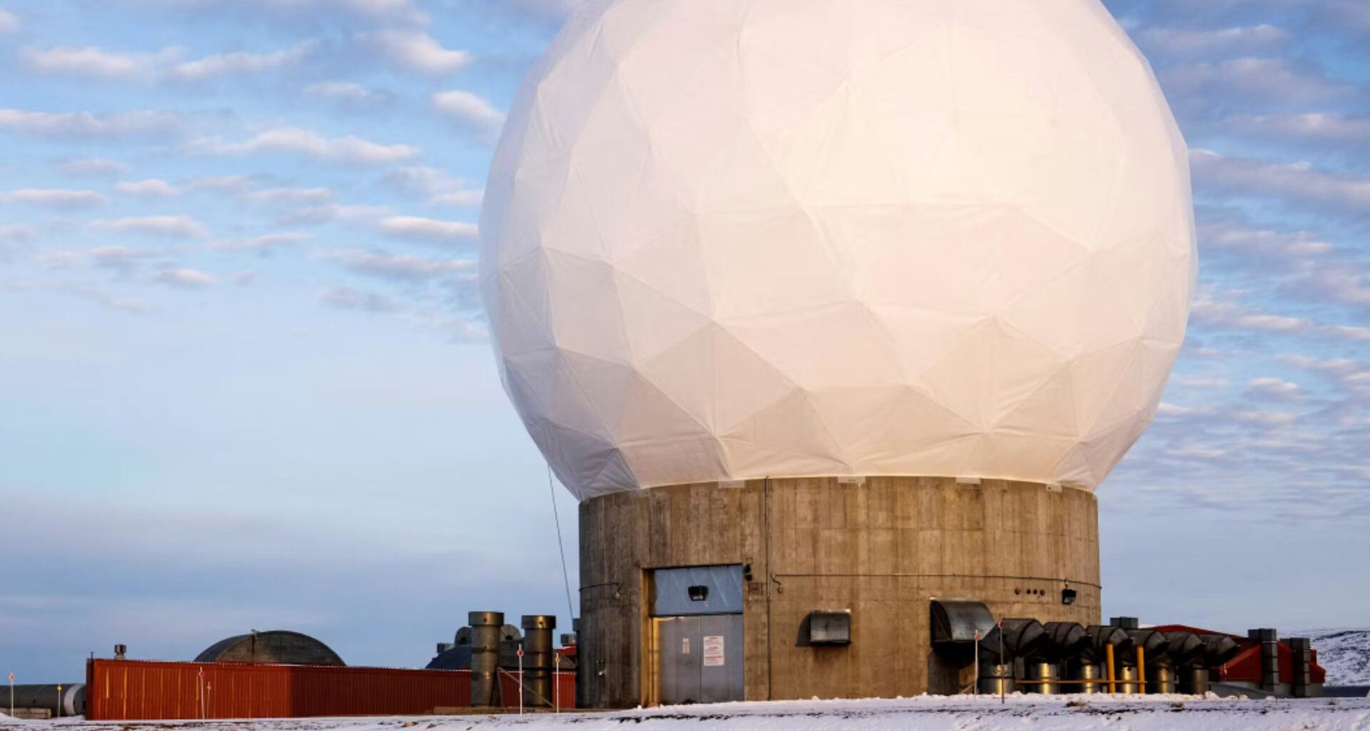 A white geodesic dome sits on top of a circular concrete base surrounded by snow under a cloudy blue sky