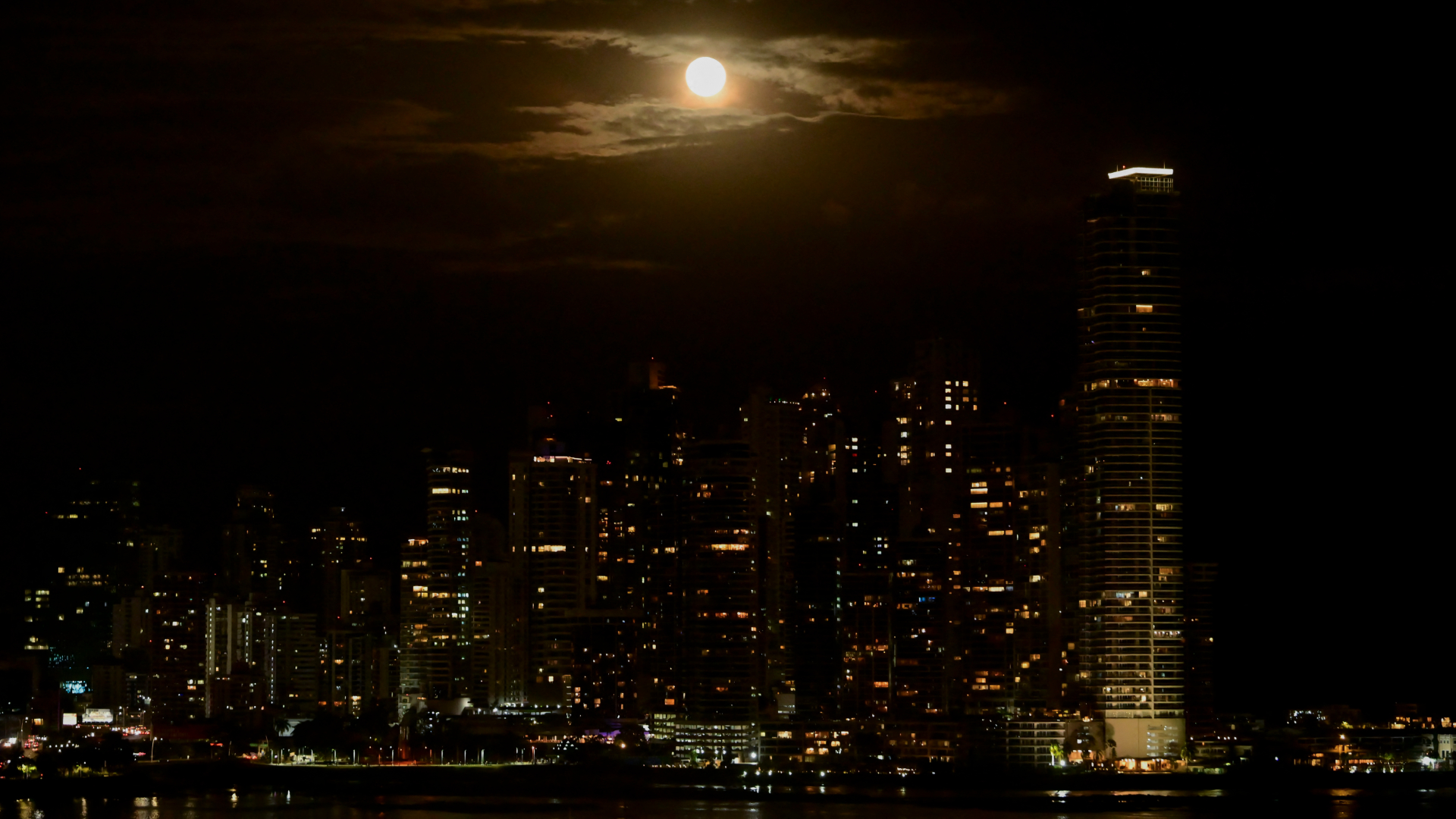 A full moon is pictured glowing brightly in a cloudy night sky above a city skyline bristling with skyscrapers.