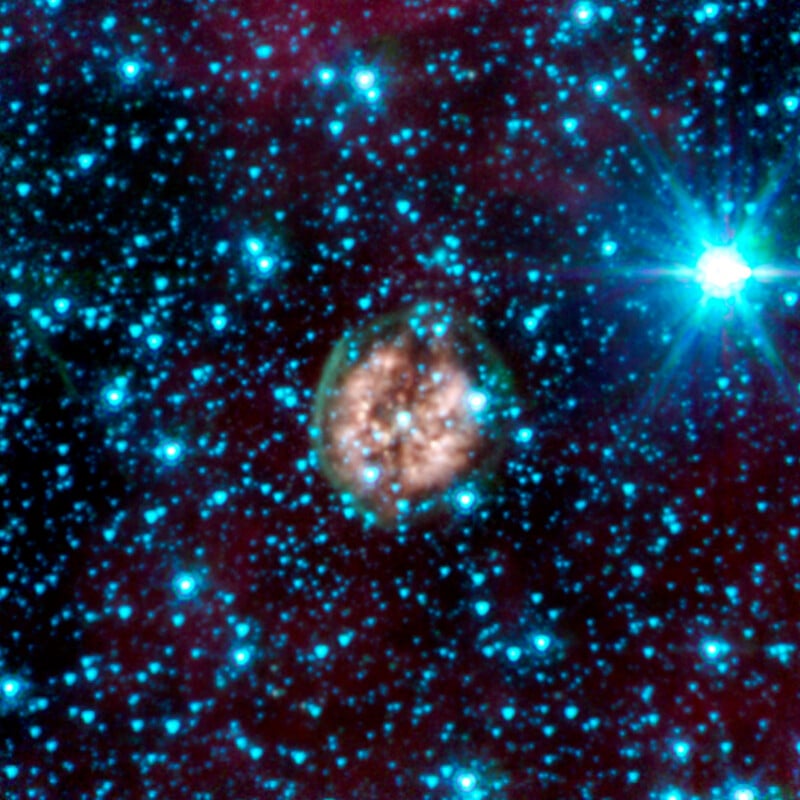 A glowing, circular nebula surrounded by numerous bright blue stars against a dark space background, with one star shining particularly brightly to the right of the nebula.