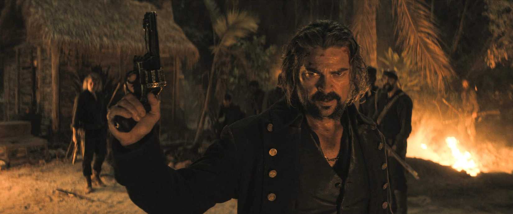 Karl Urban, playing a murderous pirate, brandishes a pistol in medium close-up in front of an evening fire in a scene from The Bluff