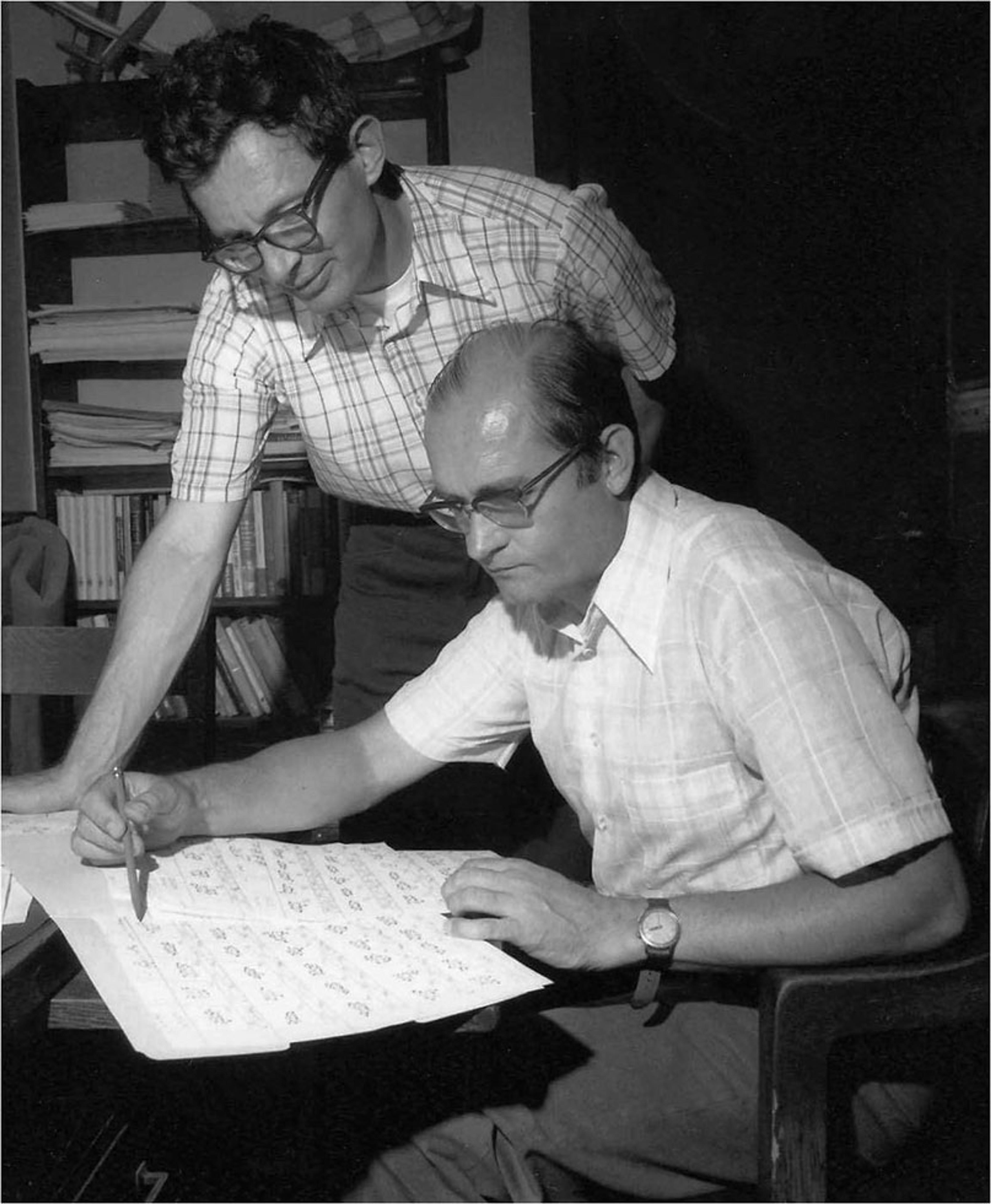 Black and white photo of two men reviewing a document at a desk in an office setting.