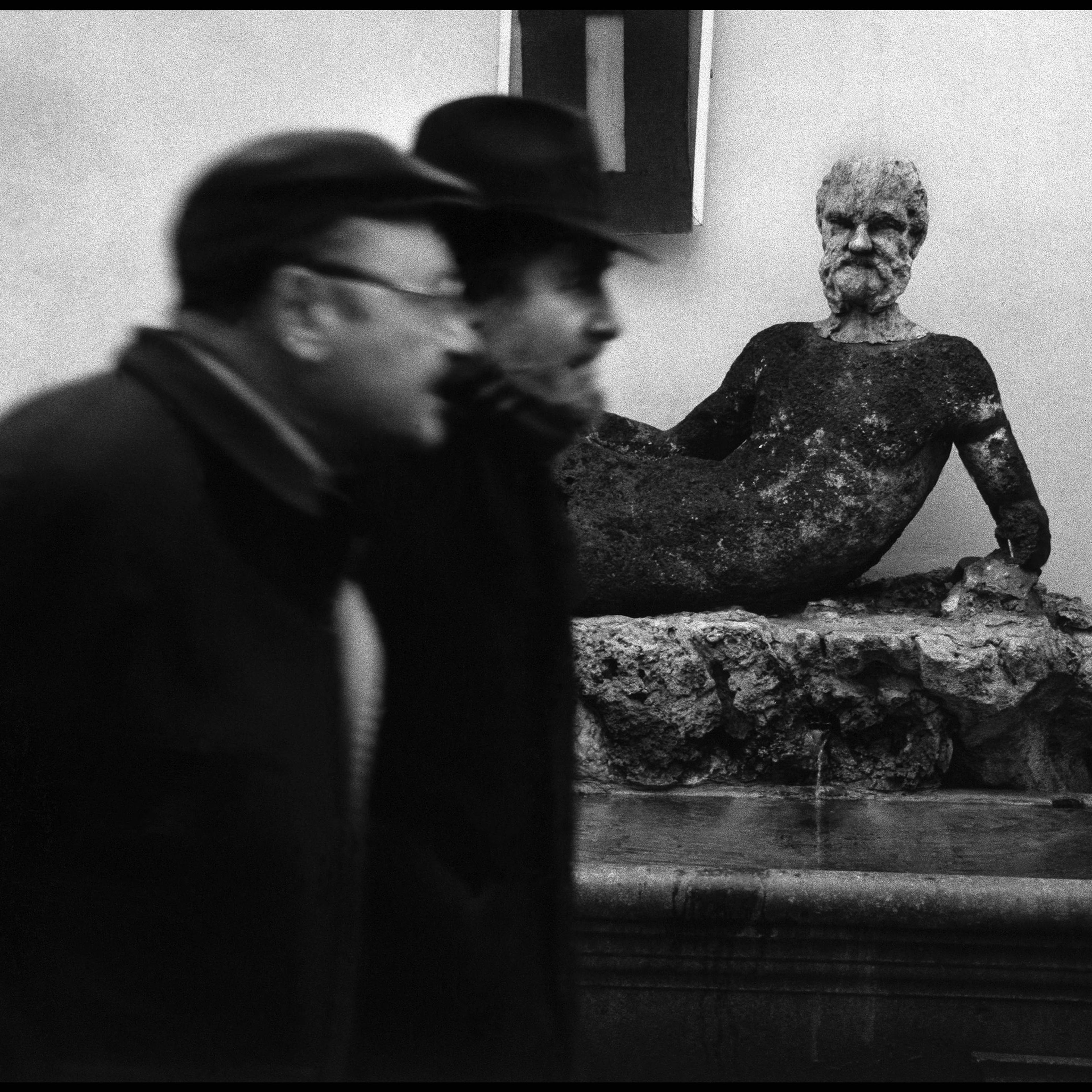 Black and white photo of two blurred men passing a weathered statue of a reclining man in a courtyard setting.