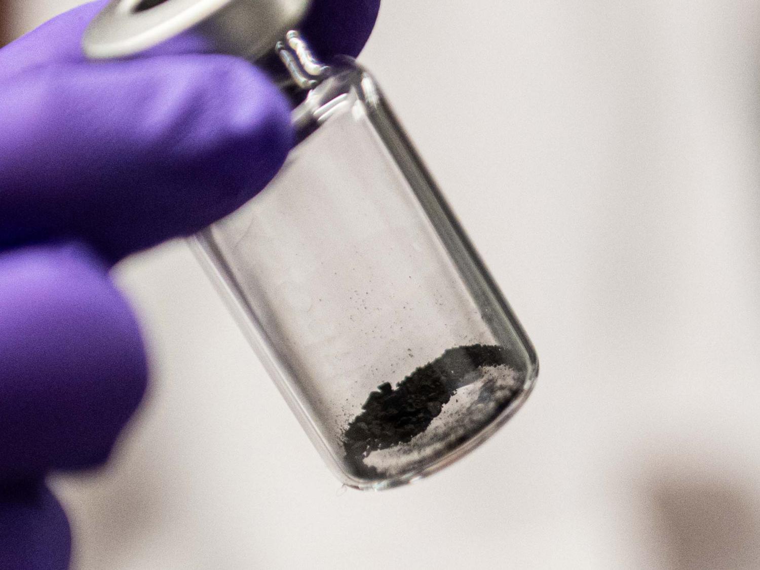 A purple gloved hand holding a clear tube with a little bit of dark gray dust within.