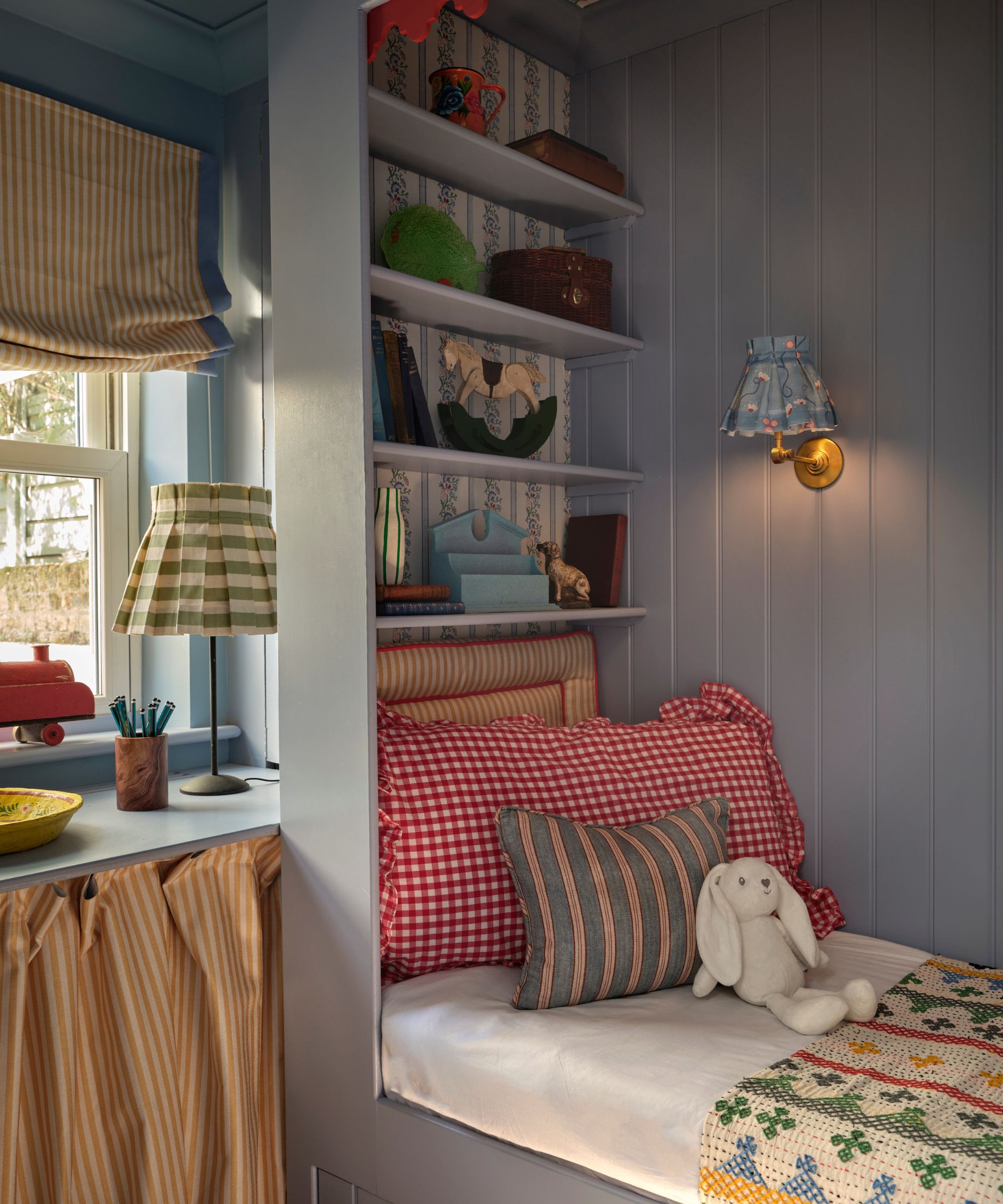 A cozy built-in sleeping nook with light blue wood-paneled walls, red gingham pillows, and a striped yellow roman shade. Built-in shelves above the bed hold toys and books.