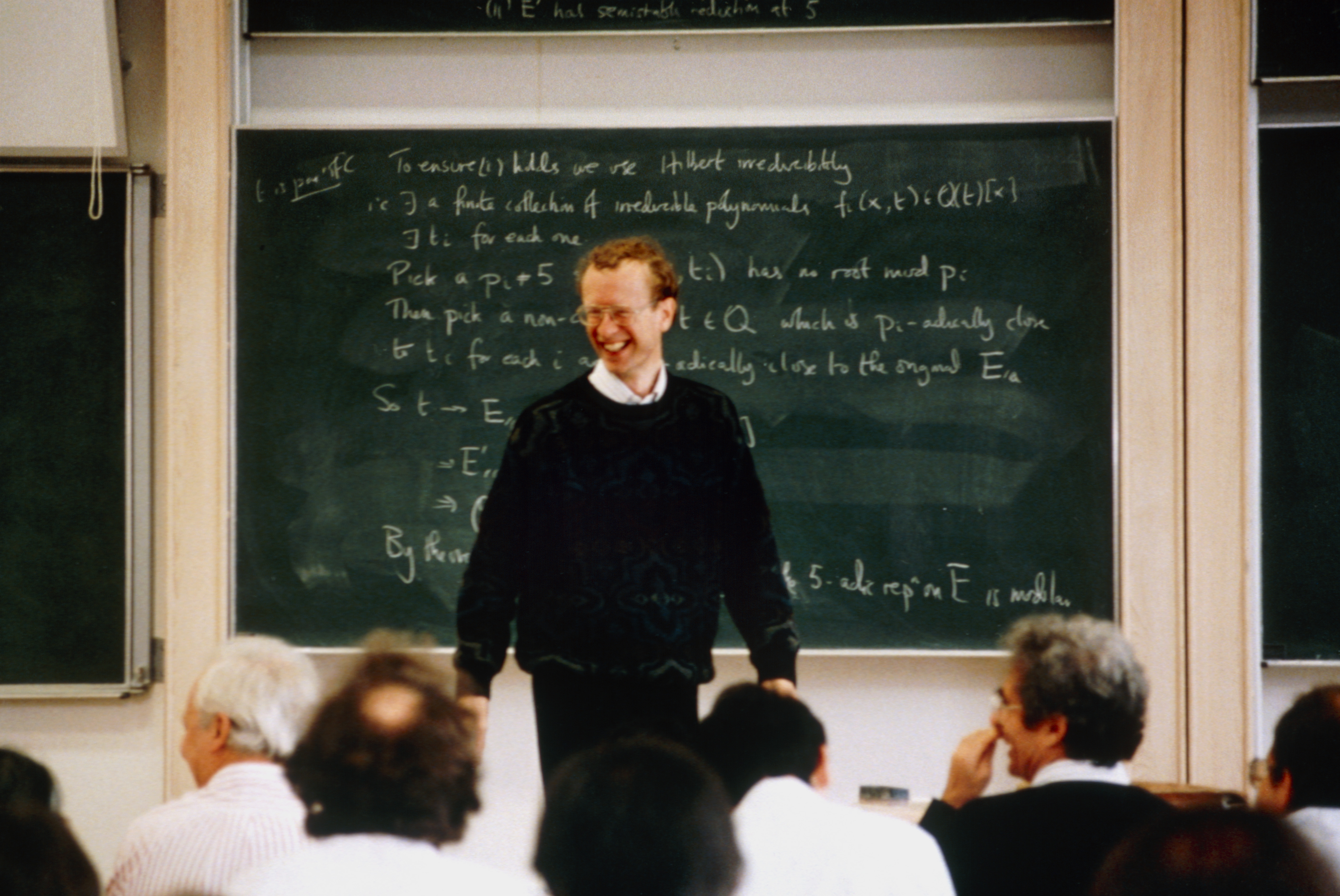 A man with curly brown hair and wireframe glasses wearing a black sweater stands in front of a green chalkboard with equations on it written in white scrawl with a seated crowd in front of him