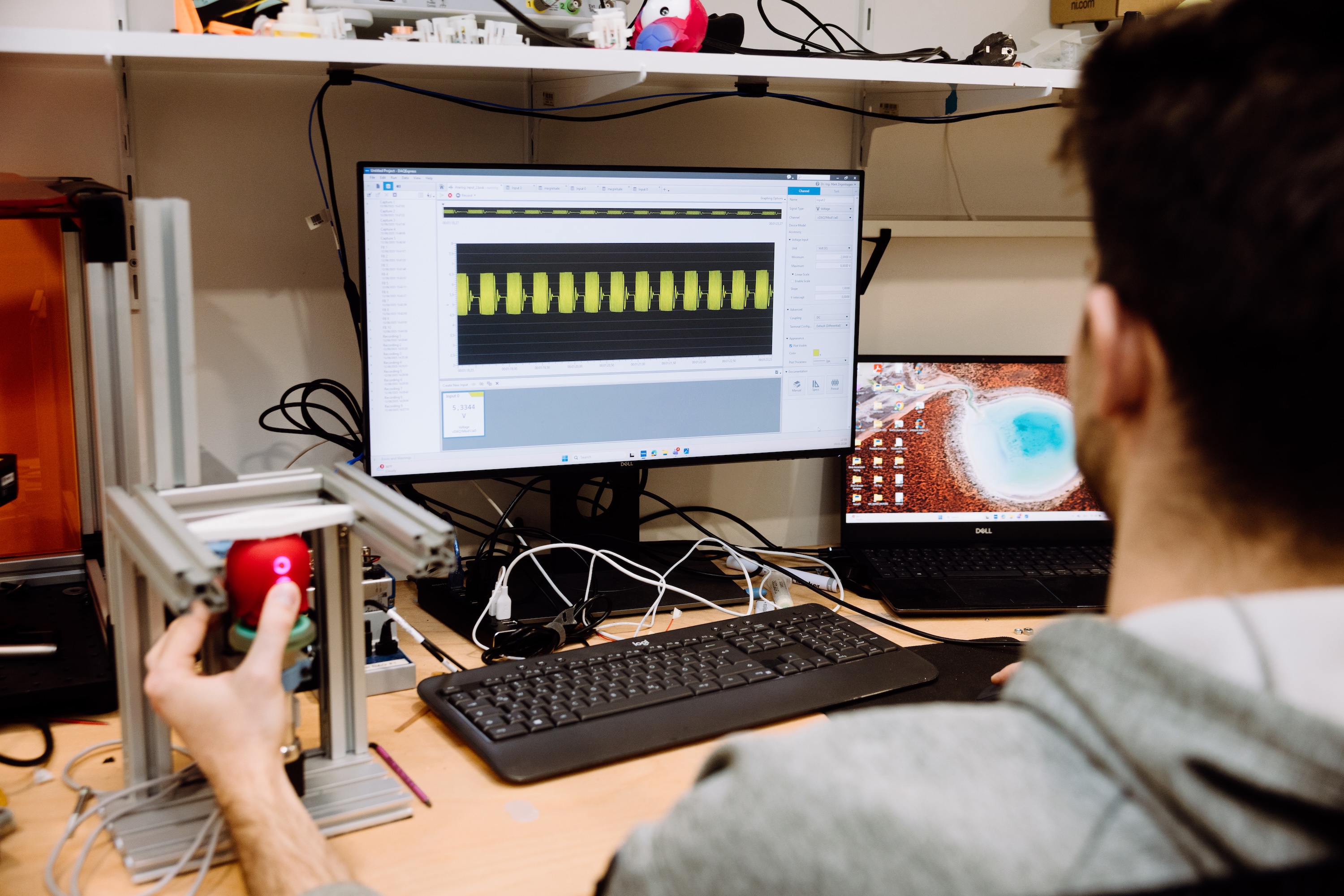 A person looking at a monitor displaying electronic signals, with a red device being held in the left foreground.