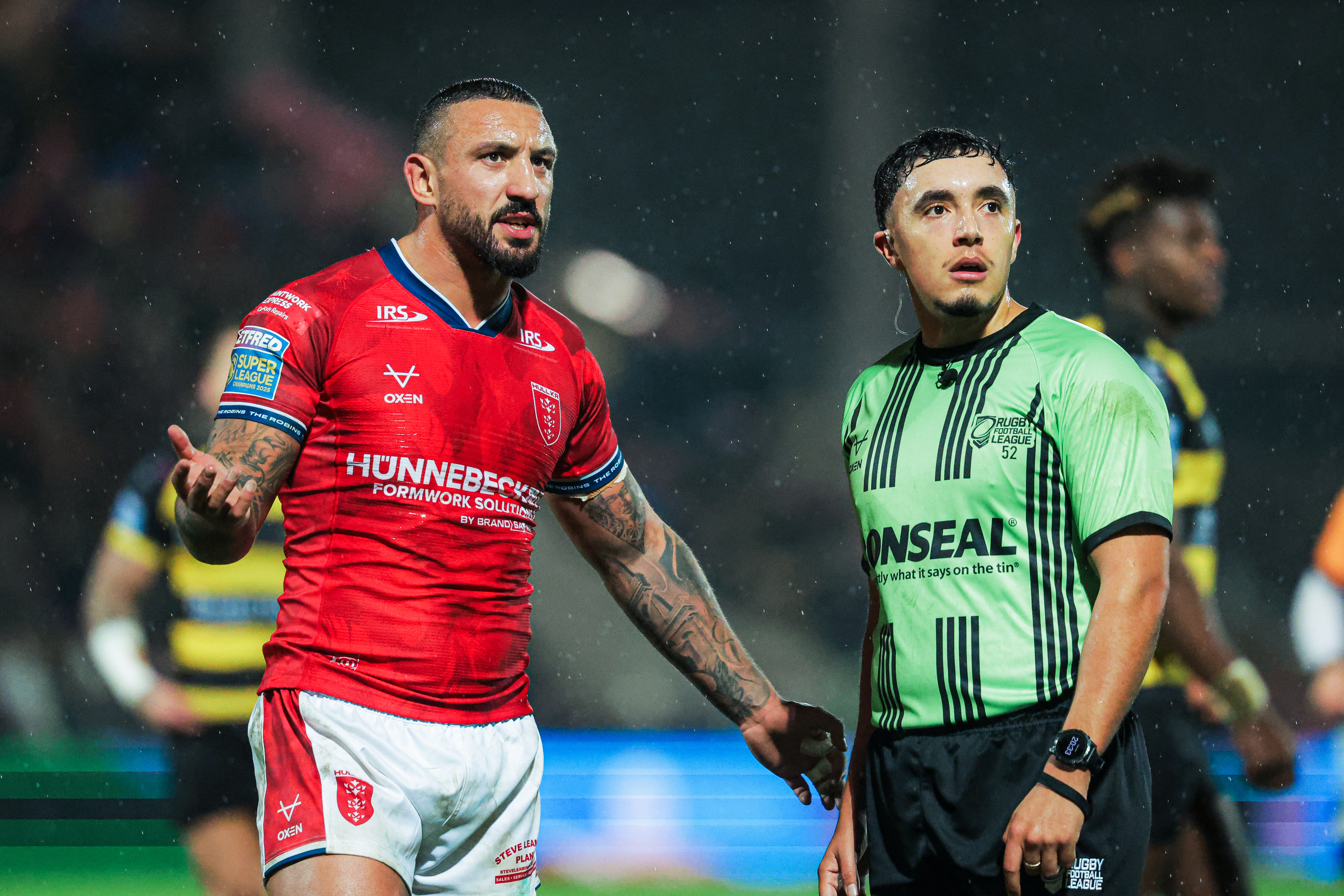 Elliot Minchella of Hull KR, in a red jersey, speaking to Referee Liam Rush, in a green and black striped jersey.