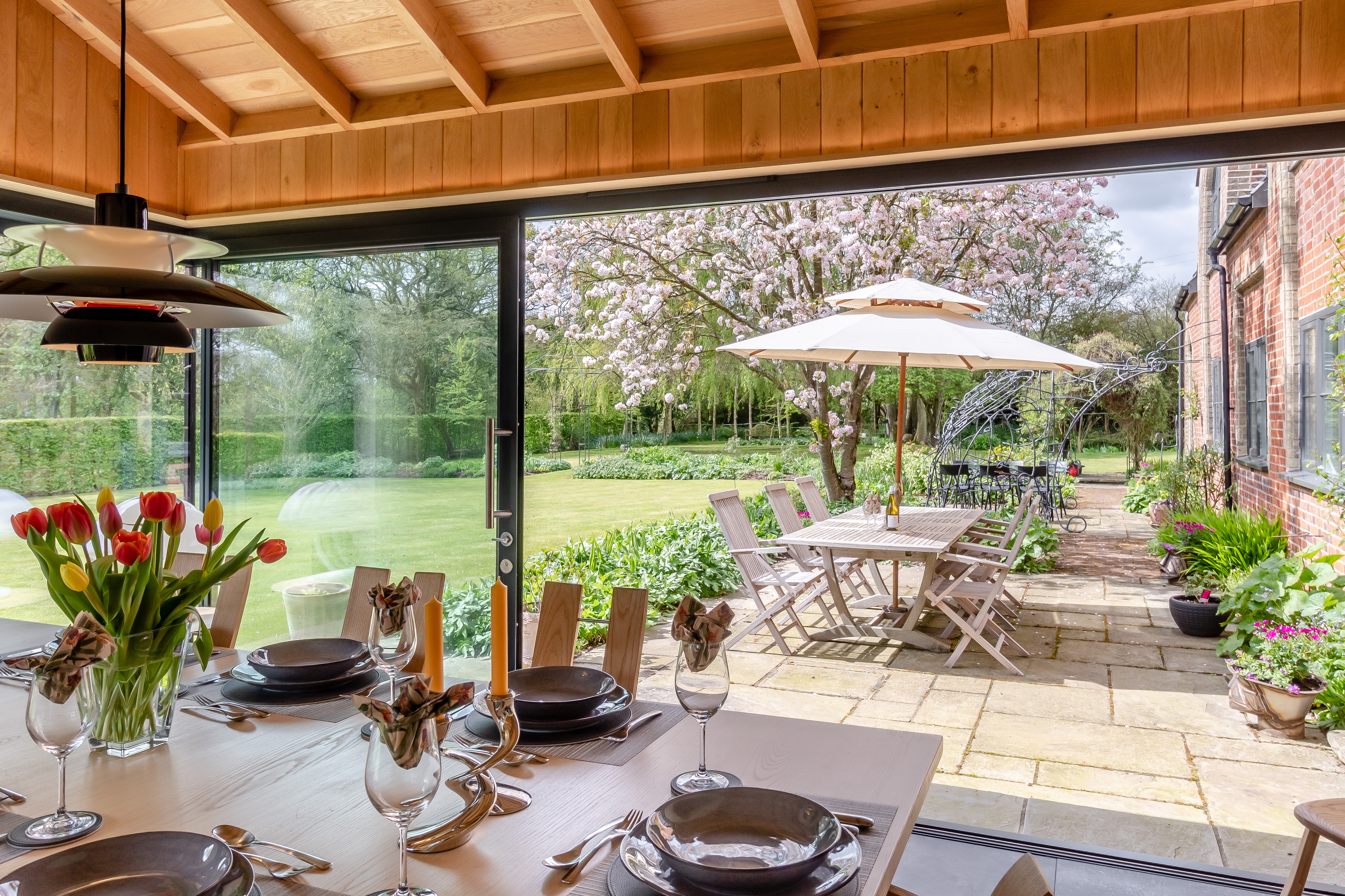 A dining room table set with plates, glasses, and flowers looking out through a glass door to a patio with outdoor seating and a blooming tree in the garden.