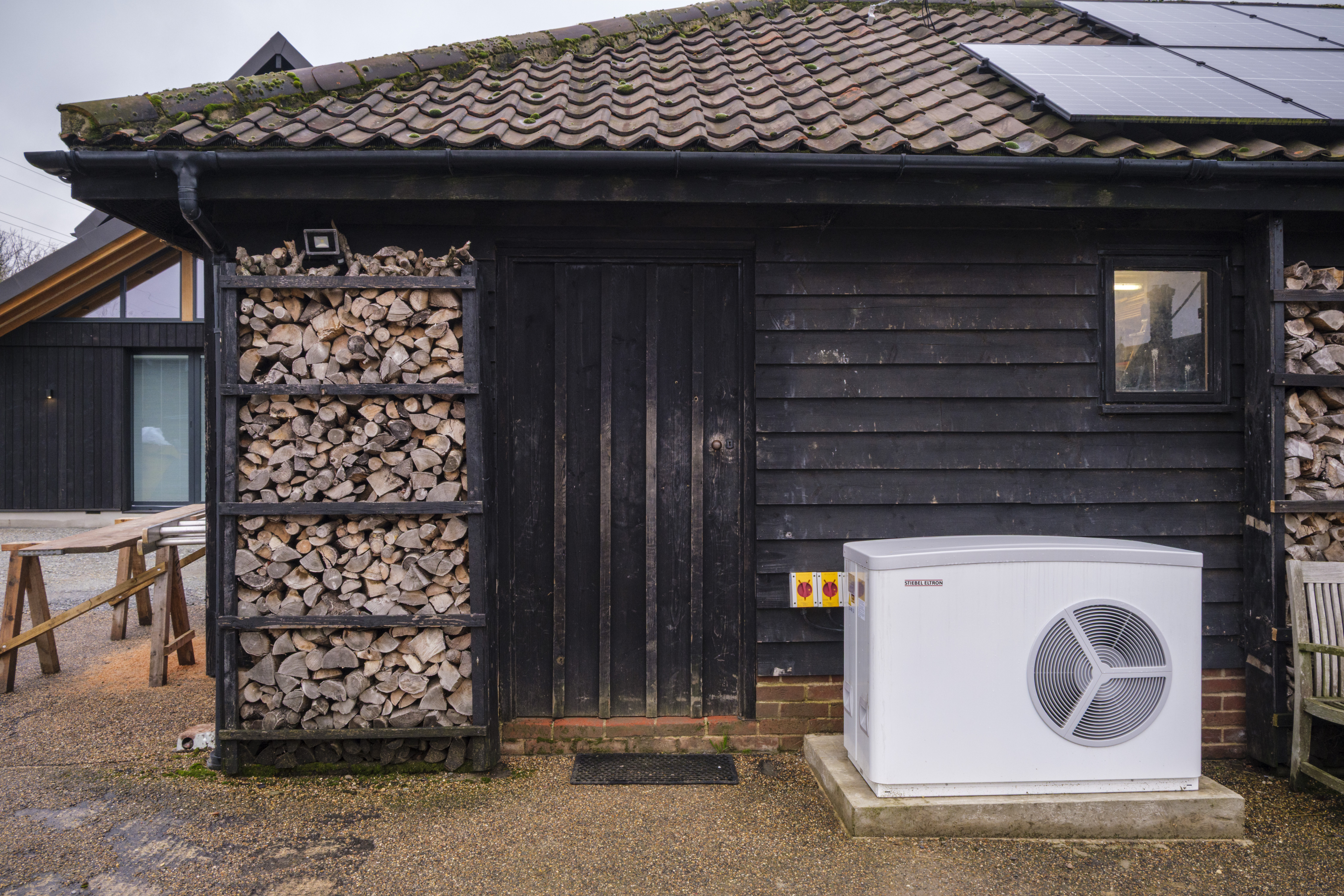 A dark-stained wooden farm building with a tiled roof, solar panels, and a modern heat pump unit.