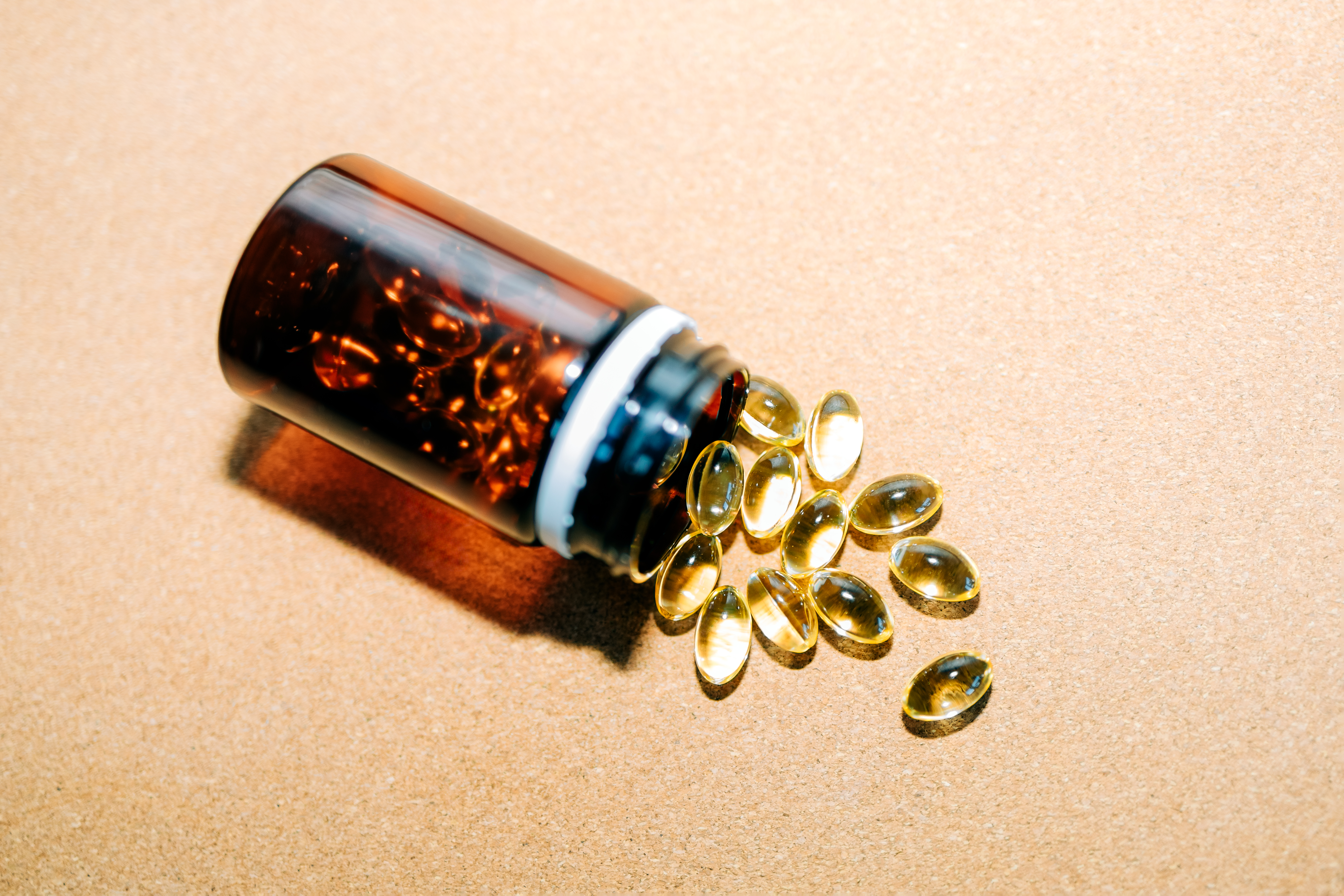 Fish oil, vitamin D, or omega-3 capsules spilling from a brown plastic bottle on a beige background.