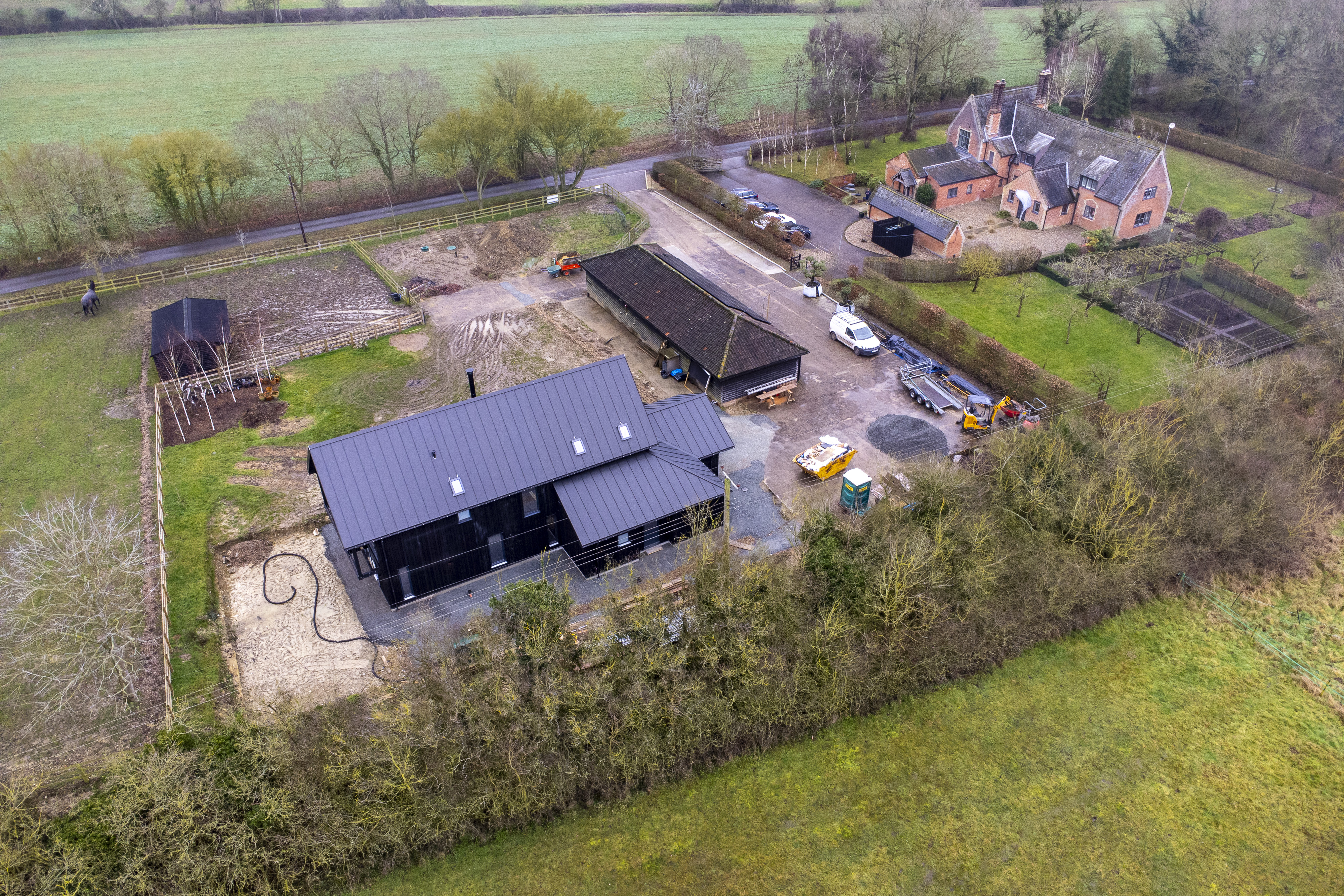 Aerial view of Becks End Farm with a new black house, a traditional brick house, outbuildings, vehicles, and surrounding fields.
