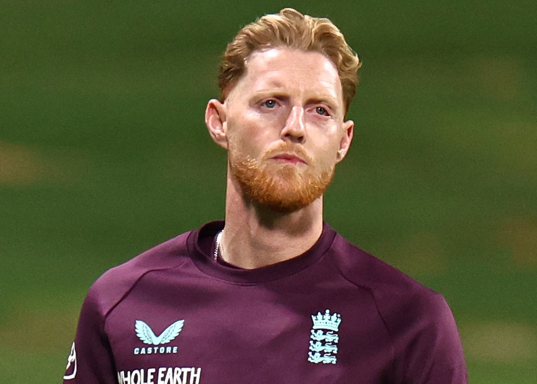 Ben Stokes walking on the cricket field, wearing a long-sleeved maroon shirt, black shorts, and white sneakers.