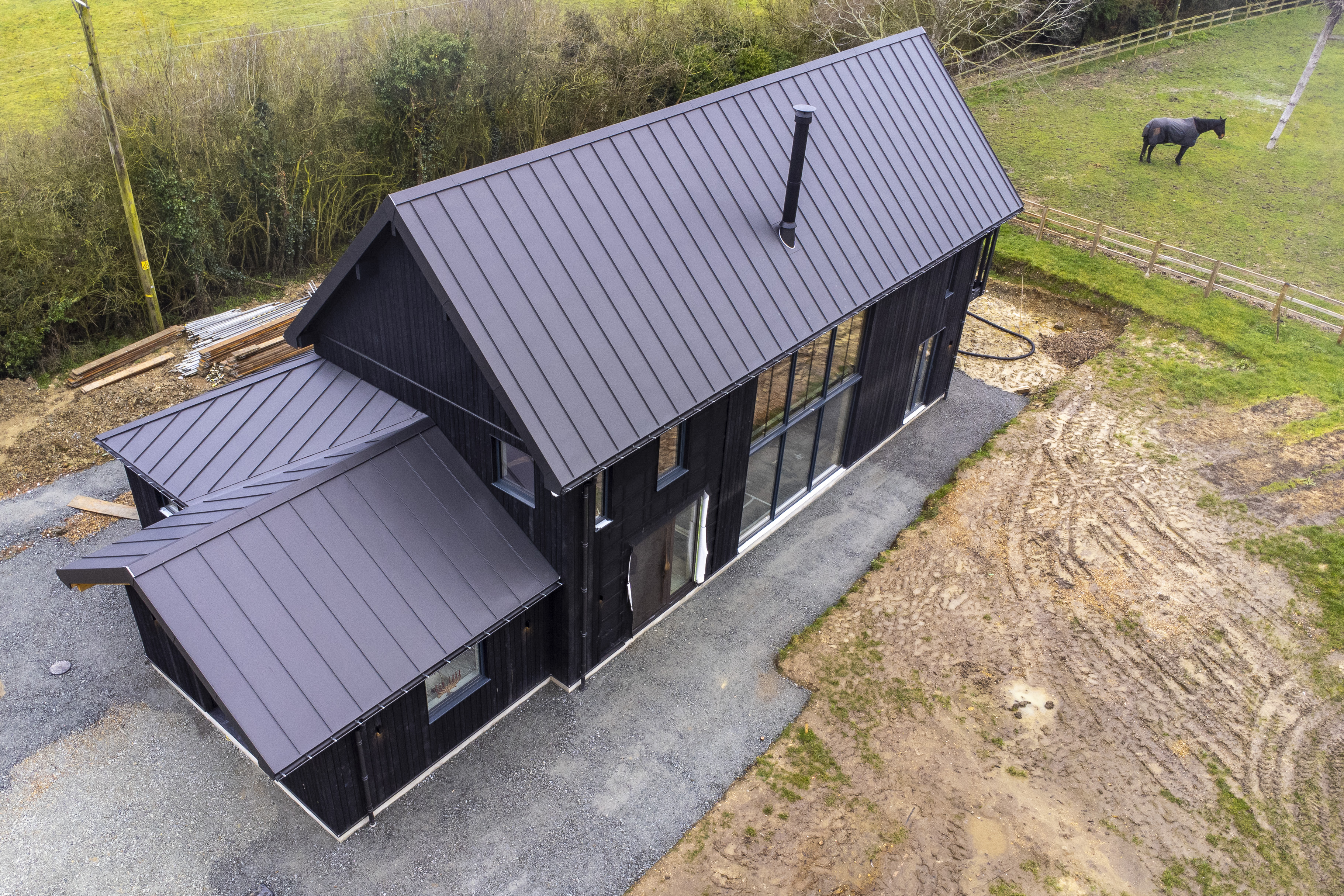 Aerial view of Becks End Farm, a black barn-style house with a dark metal roof, set next to a muddy field with a horse.