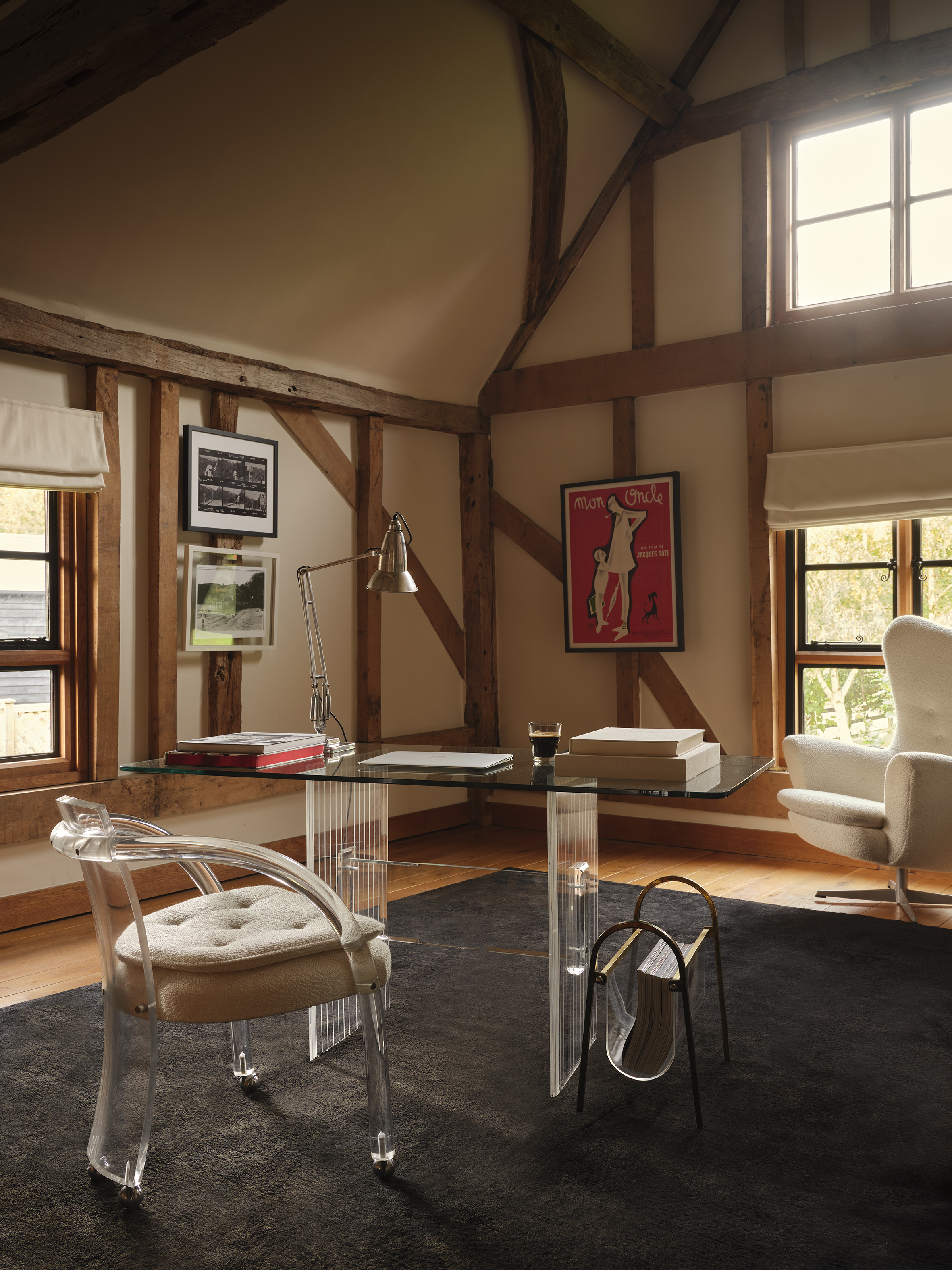 A modern home office with a glass desk, clear acrylic chair, and a fluffy white armchair next to a window.
