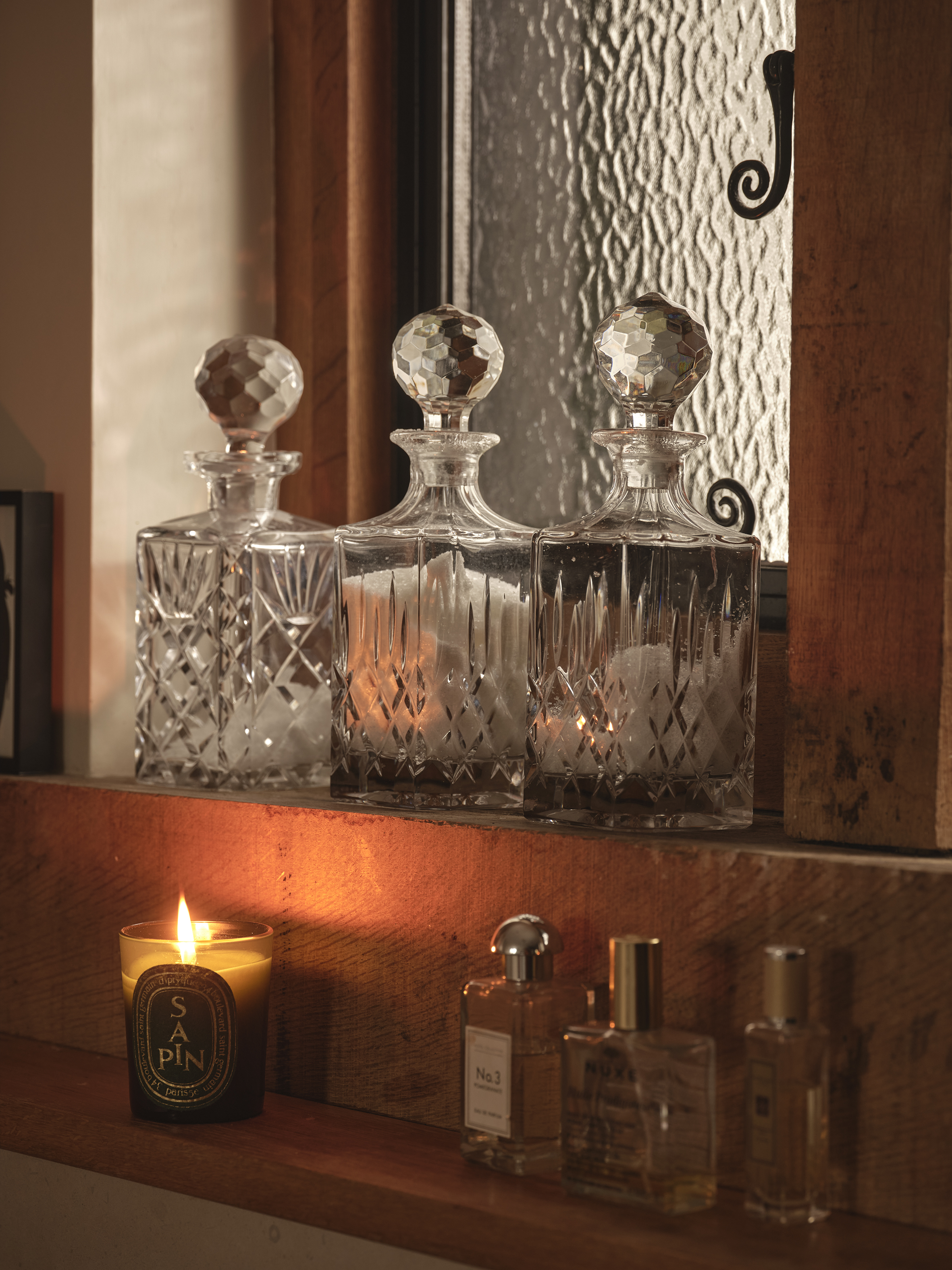 A lit candle and perfume bottles on a wooden shelf, with three crystal decanters on a higher shelf.