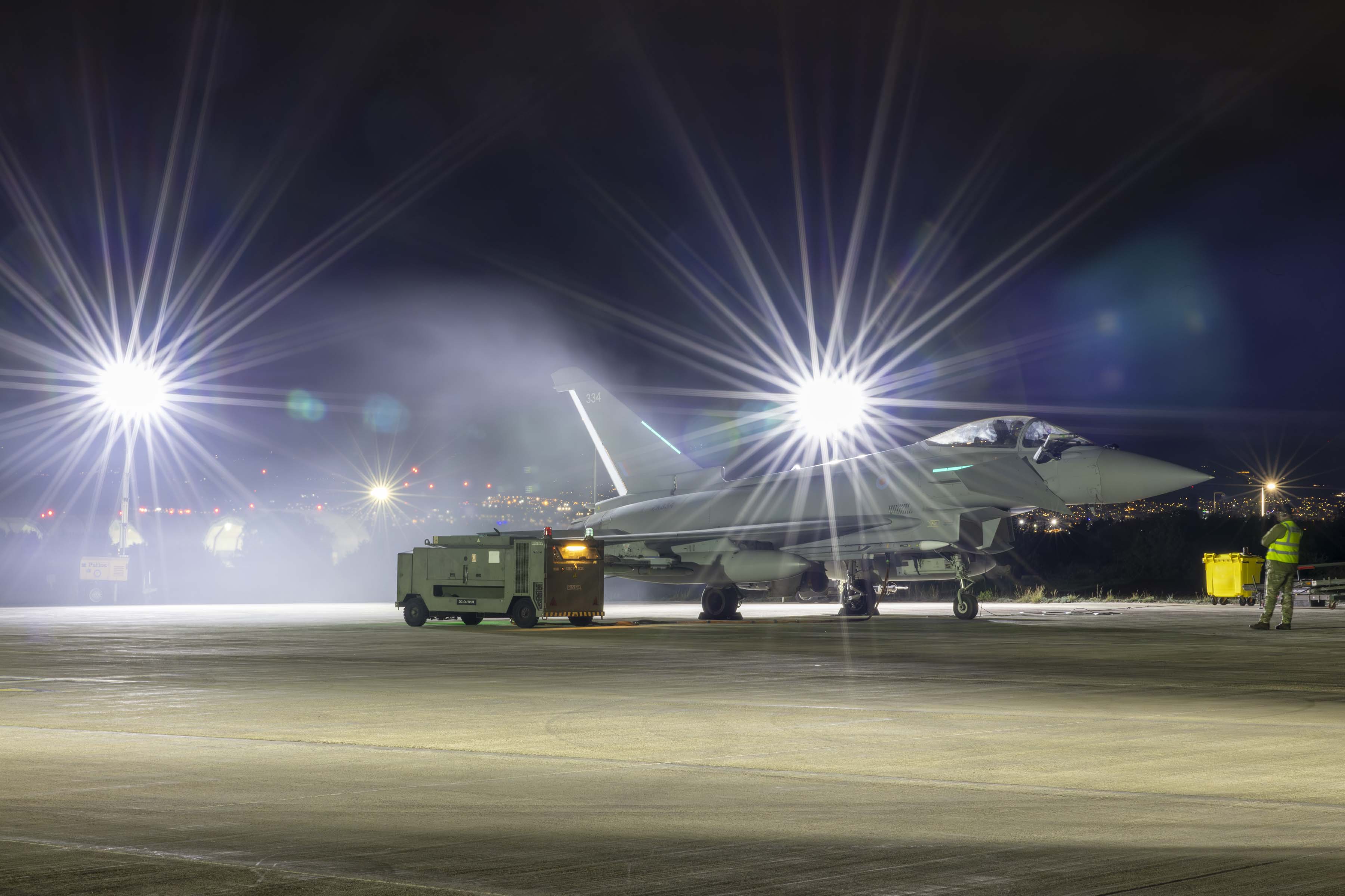 An RAF Typhoon aircraft on the tarmac at night, with two bright lights shining from the left side.