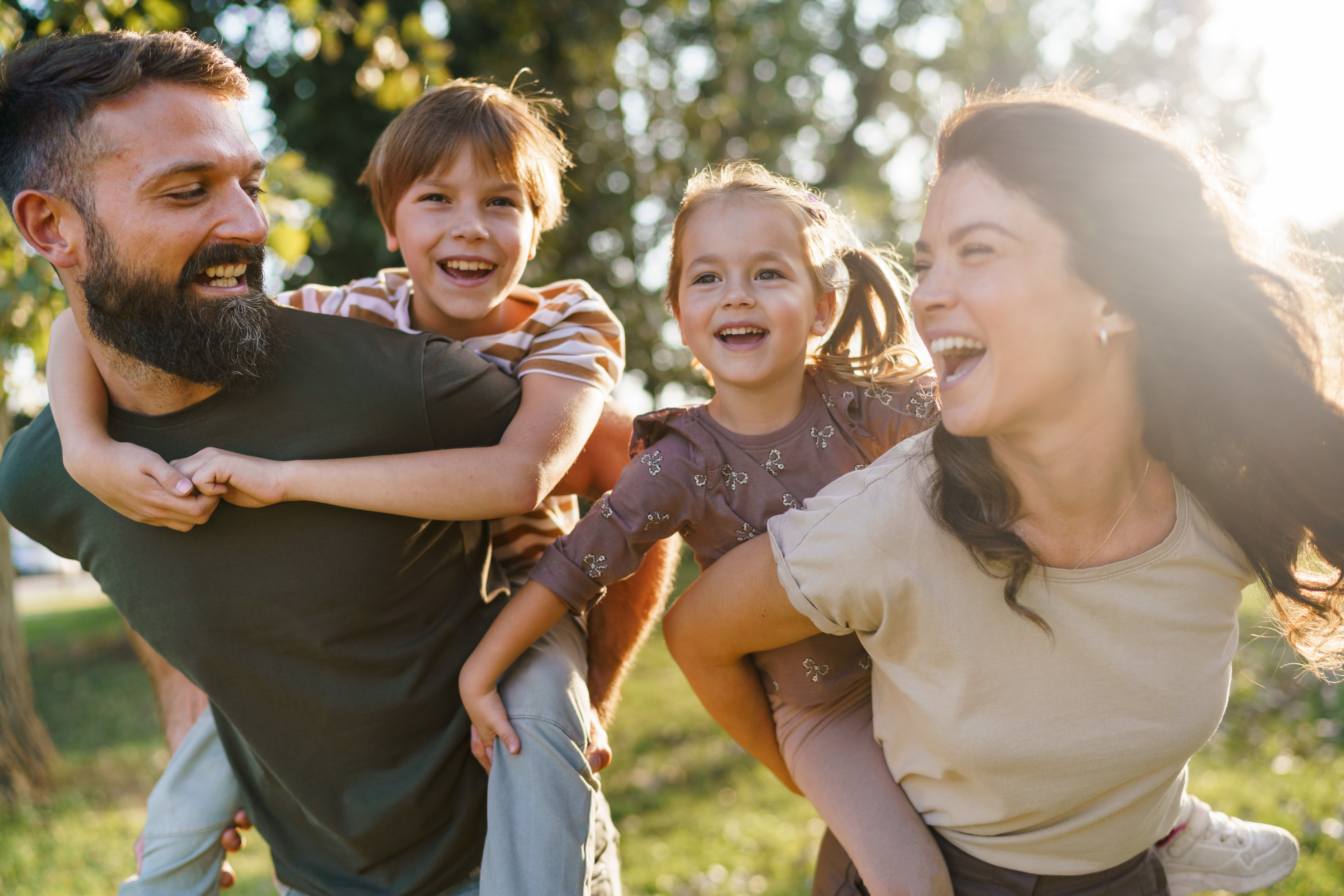 A father and mother giving piggyback rides to their son and daughter, all laughing joyfully outdoors.