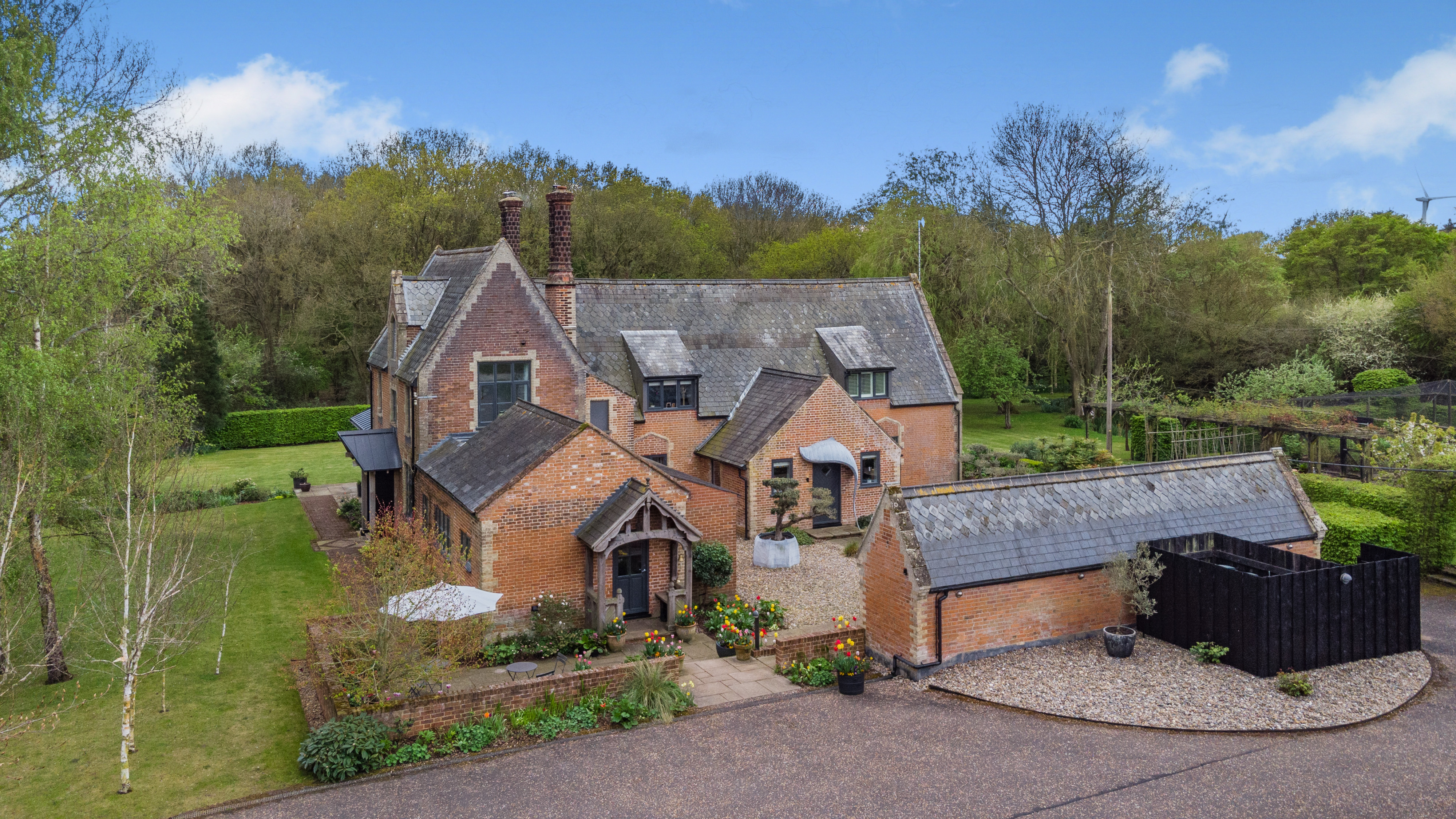 An aerial view of the Becks End Farm property, showing a large brick house with dark roofs and a black outbuilding with solar panels, surrounded by lawns and fields with horses.