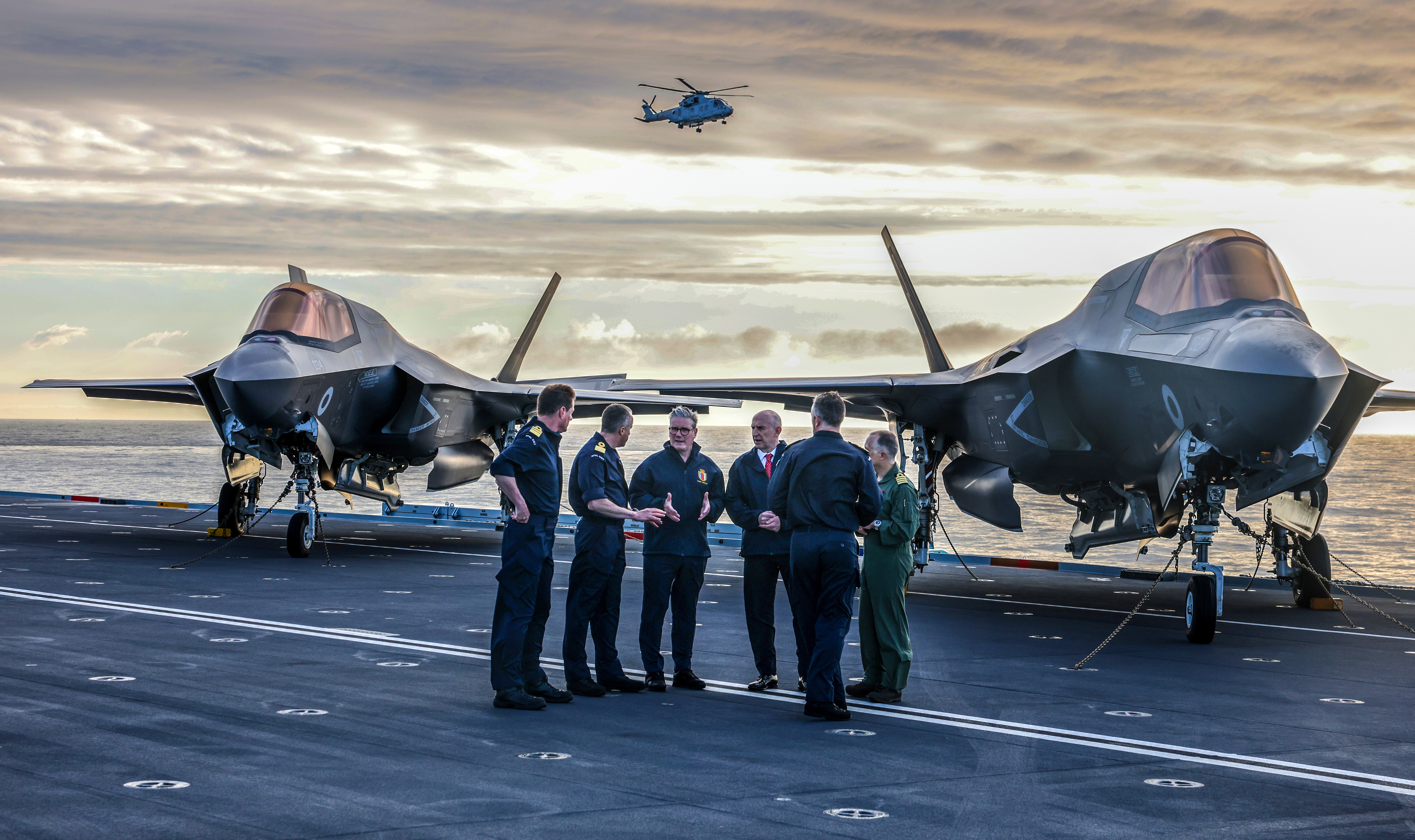 Prime Minister Keir Starmer and Defence Secretary John Healy talk with ship's officers in front of parked F35 fighter jets on an aircraft carrier.