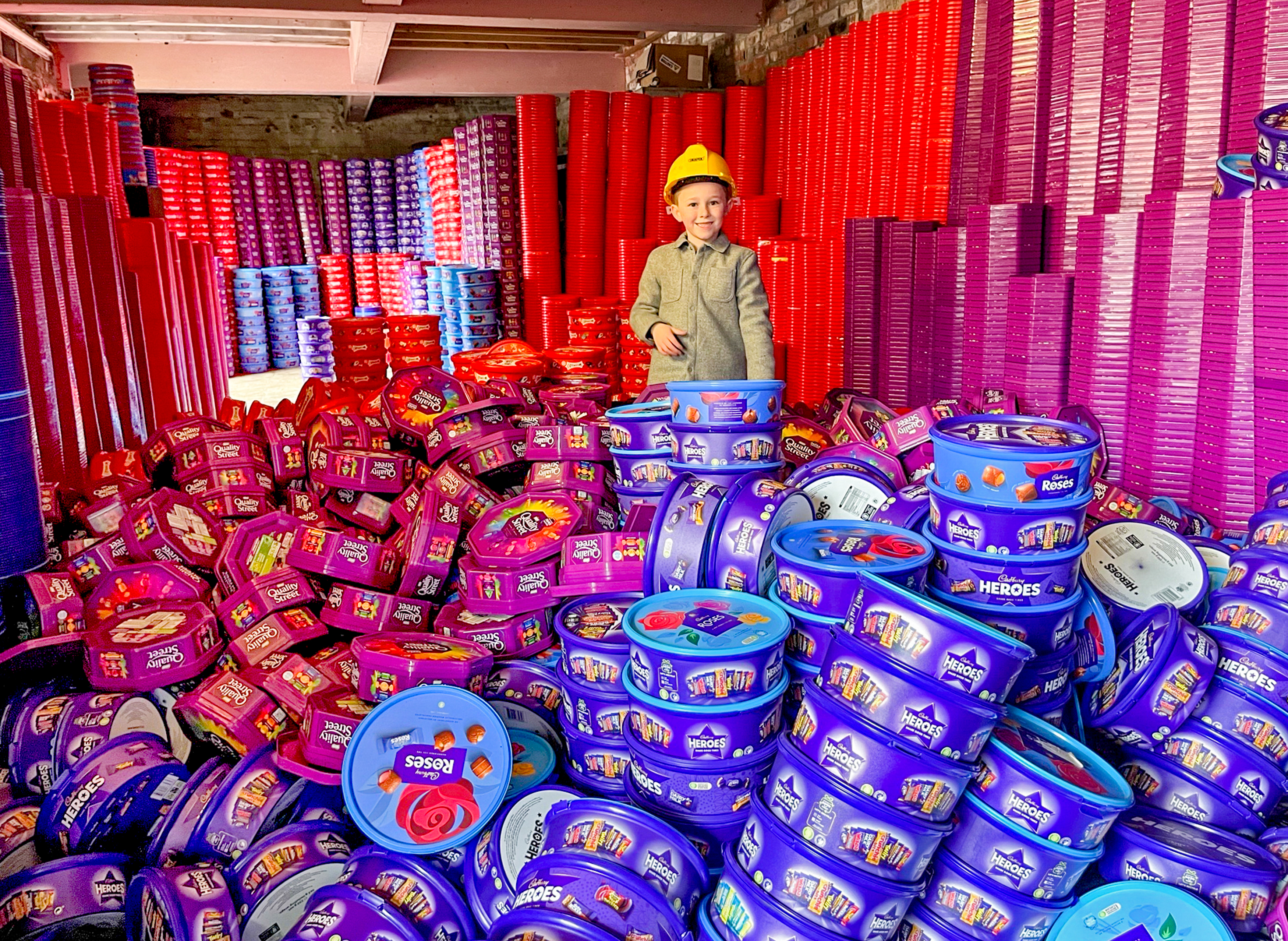A young boy in a yellow hardhat surrounded by stacks and piles of colorful plastic sweet tubs.