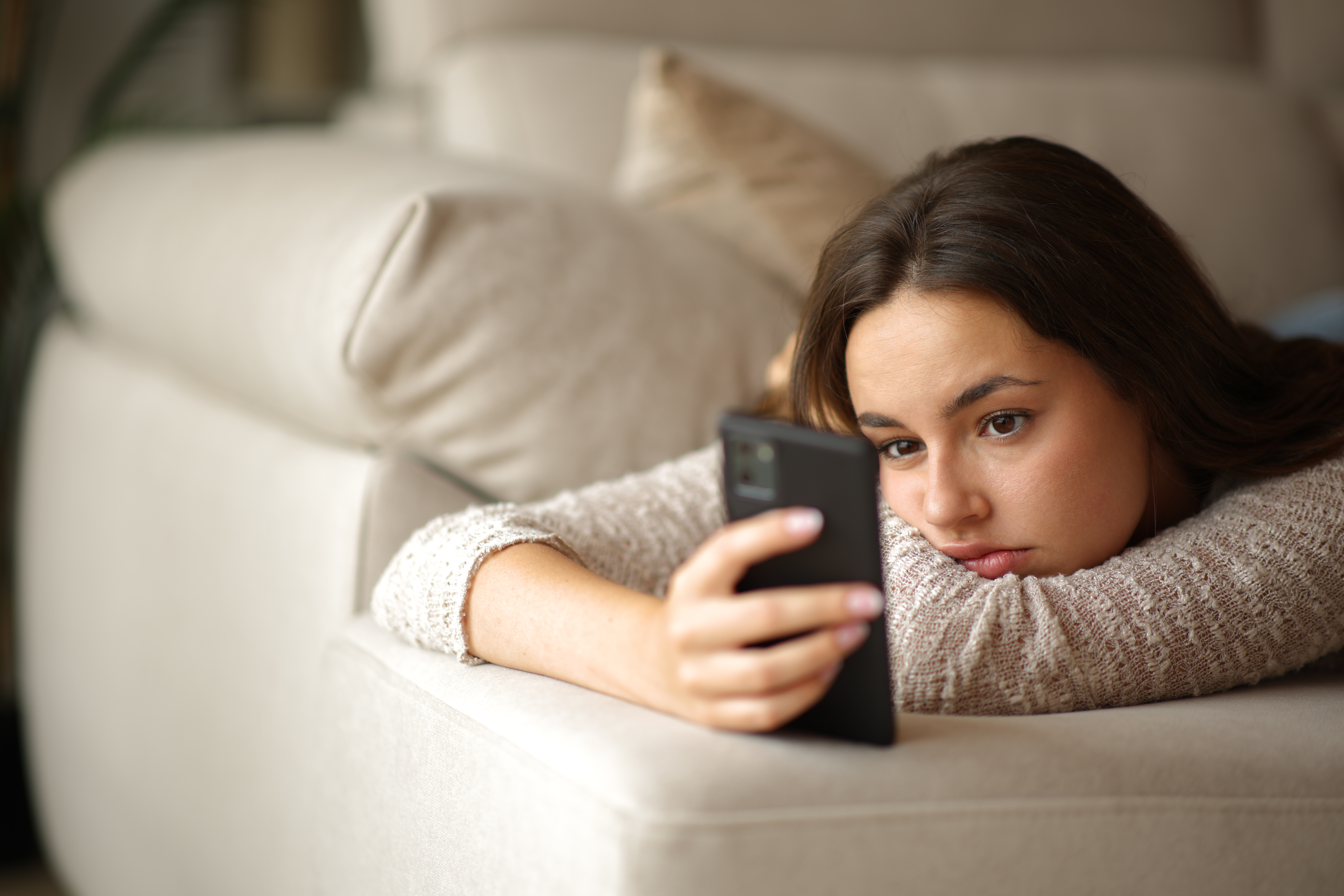 A bored woman lying on a couch, holding and looking at her smartphone.