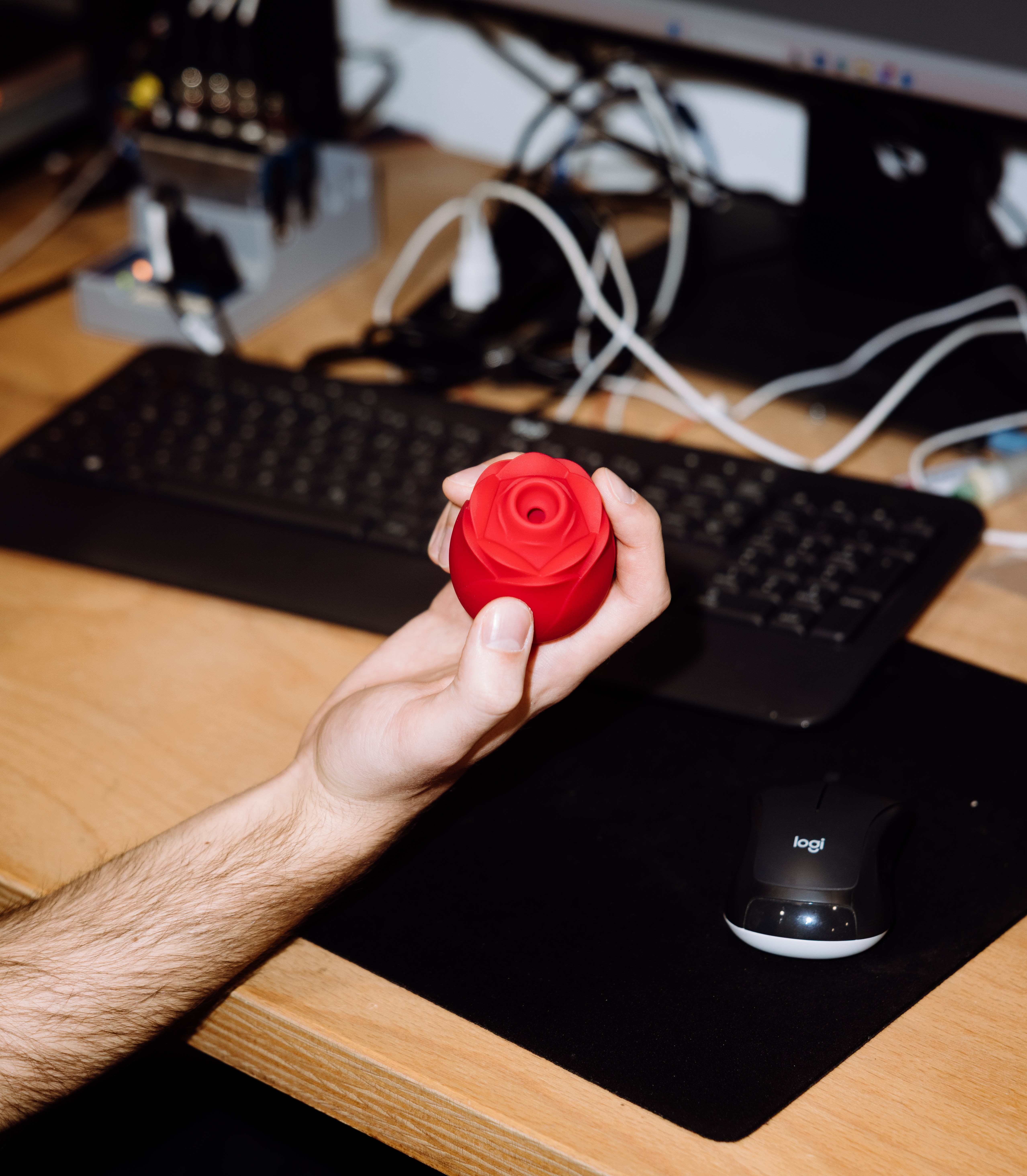 A hand holds a red rose-shaped sex toy above a desk with a keyboard and mouse.