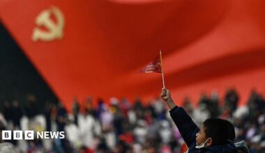 A little boy raises a small flag at the plaza of the Museum of the Communist Party of China in Beijing. The foreground is blurred, but one can make out a sculptural flag of the Communist Party in the background