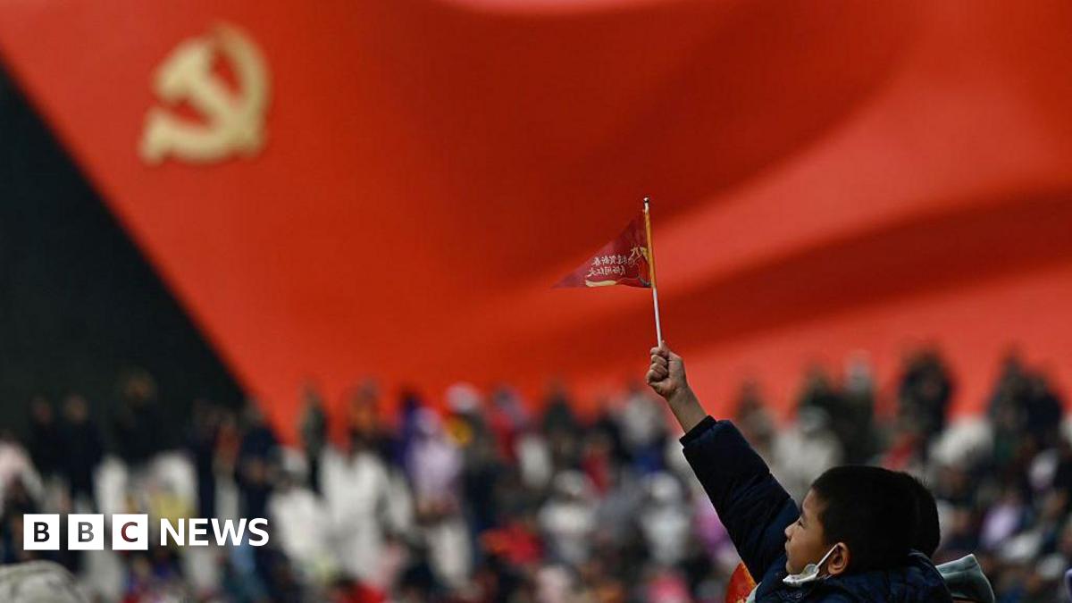 A little boy raises a small flag at the plaza of the Museum of the Communist Party of China in Beijing. The foreground is blurred, but one can make out a sculptural flag of the Communist Party in the background