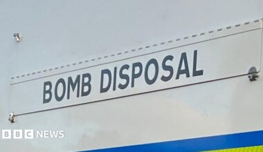 A small robot with rubber tracks, mounted cameras, coiled cables, and an articulated robotic arm positioned forward.