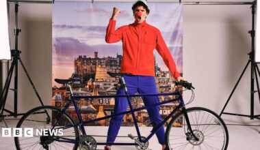 Greg James standing behind a tandem bicycle wearing blue trousers, a red top baseball cap and a red nose on his nose.