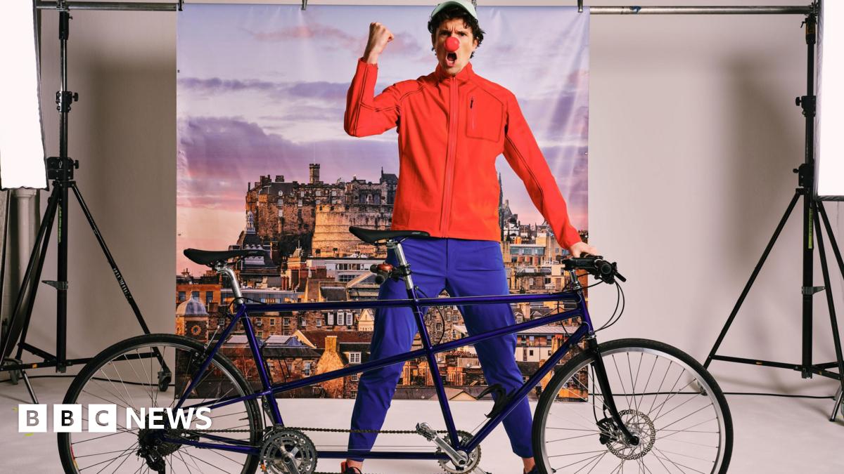 Greg James standing behind a tandem bicycle wearing blue trousers, a red top baseball cap and a red nose on his nose.