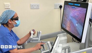 A female consultant in blue scrubs, glasses and face mask operates a machine which carries out robotic hysterectomies.