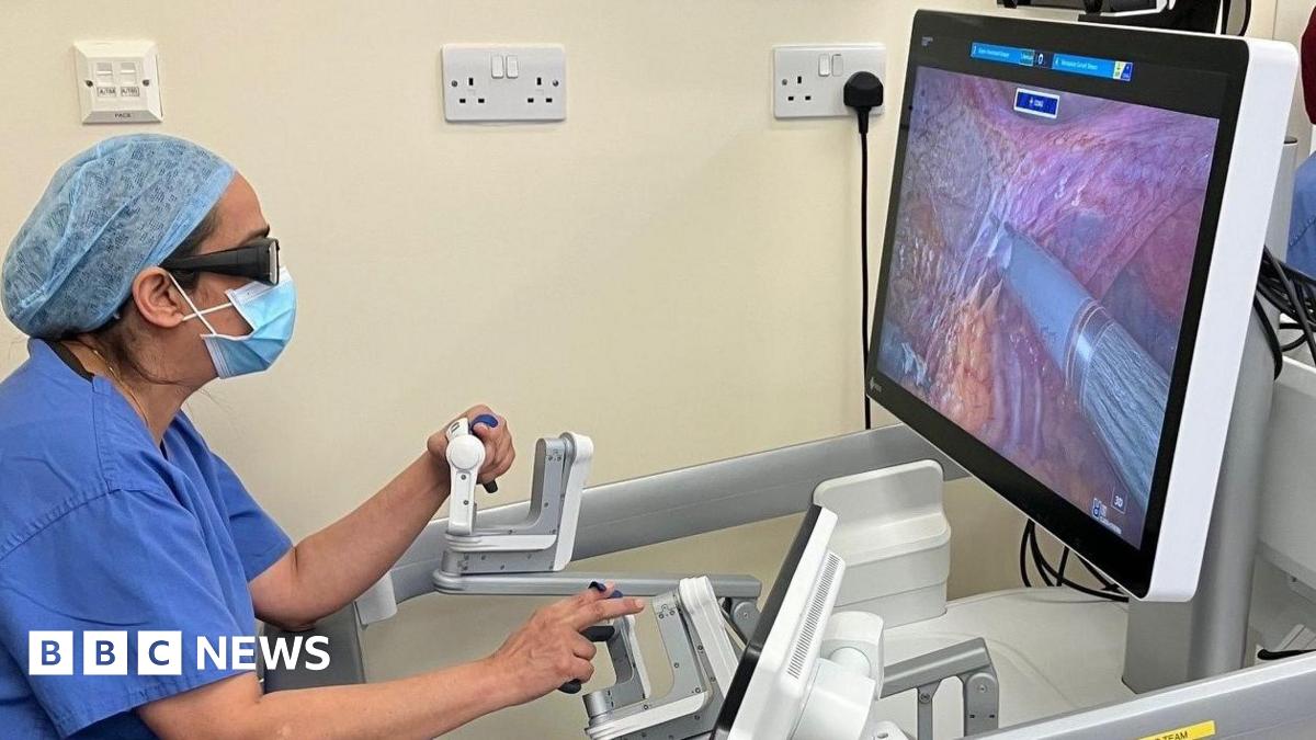 A female consultant in blue scrubs, glasses and face mask operates a machine which carries out robotic hysterectomies.